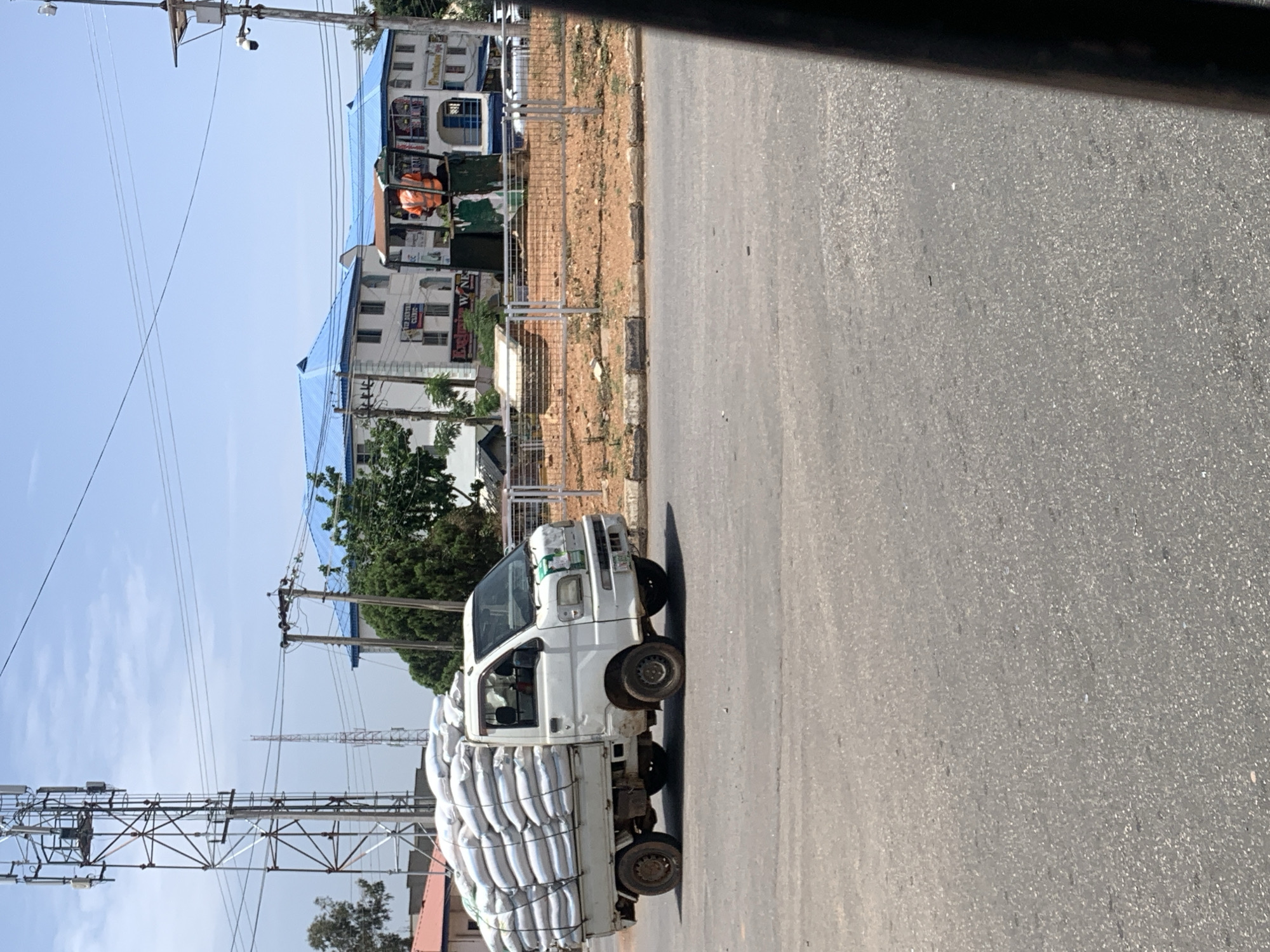A white truck heavily laden with sacks travels on an asphalt road in Jos, Nigeria. The truck's condition suggests age and use. The road borders a built-up area, with multi-story buildings visible. A communication tower is present on the side of the road opposite the buildings. The day appears clear.