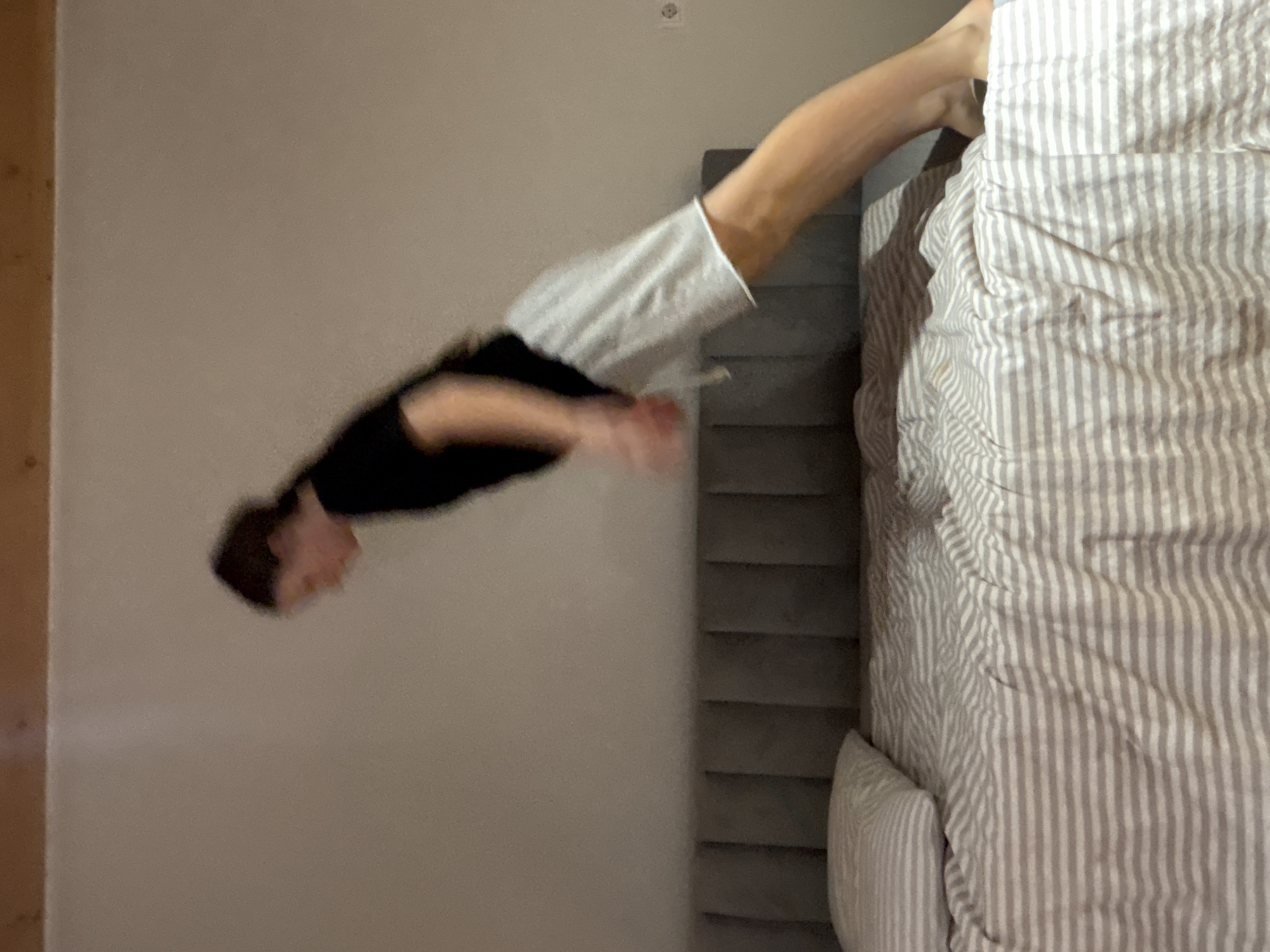 A blurred image shows a person in a black top and white pants seemingly diving onto a bed with striped sheets and a gray headboard. The background consists of a light gray wall with a possible electrical outlet near the top and a wooden door frame on the left side. The location is assumed to be Chaikovsky, Russia. The subject is blurred, with the head near the left of the frame and the hand extended.