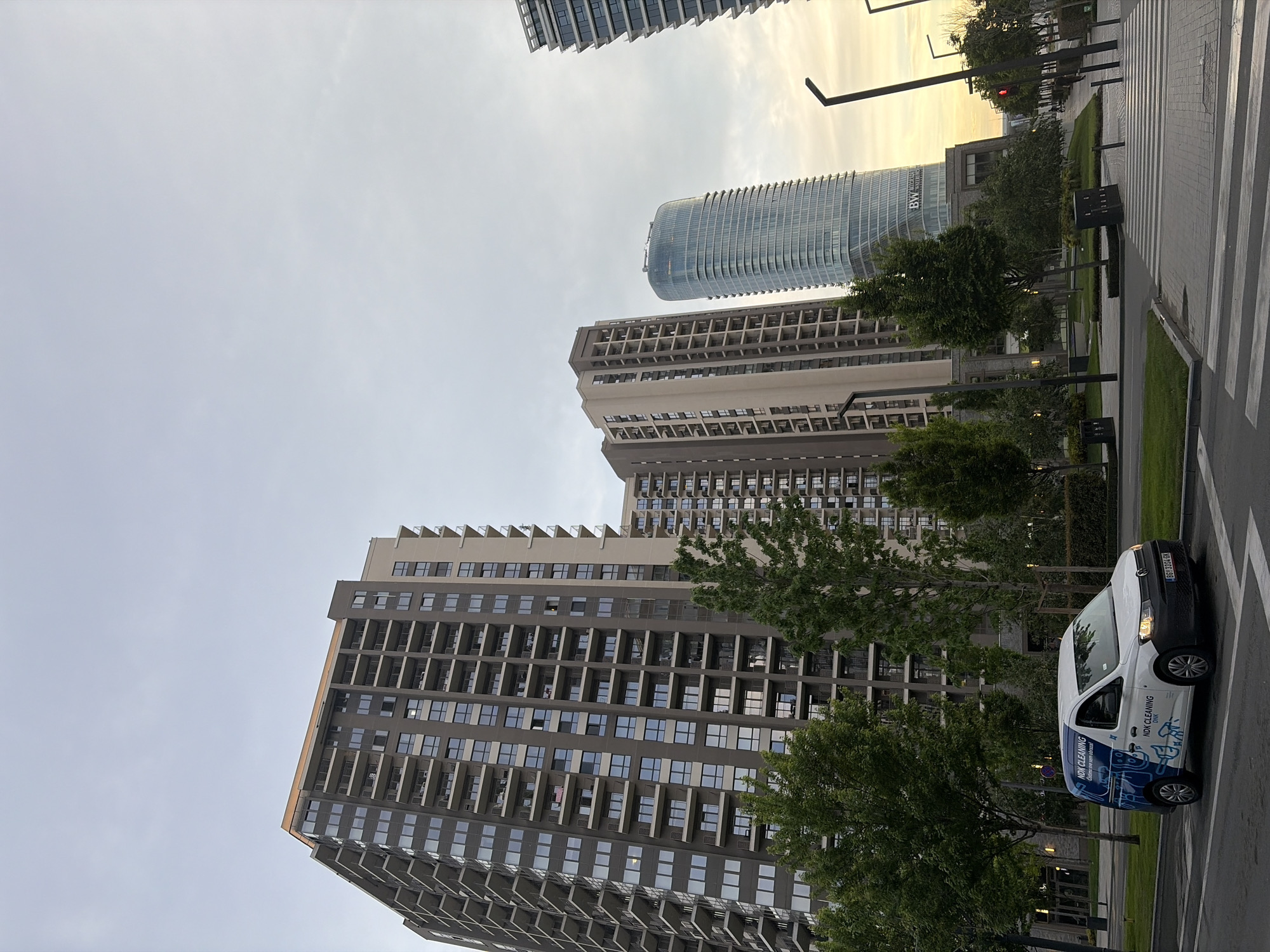 The image depicts a street scene in Belgrade, Serbia, specifically the Belgrade Waterfront area, characterized by modern high-rise buildings. The buildings are a mix of residential and commercial structures with rectangular shapes and numerous windows. The sky is overcast, possibly indicating early evening or morning. A cleaning service van is parked on the street next to a crosswalk. The van is labeled "NDK Cleaning" with contact information. The street is clean and well-maintained. Pedestrian crosswalks are visible. The environment suggests a modern urban landscape with landscaped areas. Trees are dispersed throughout the scene. A stoplight is visible in the background. There are no people visible.
