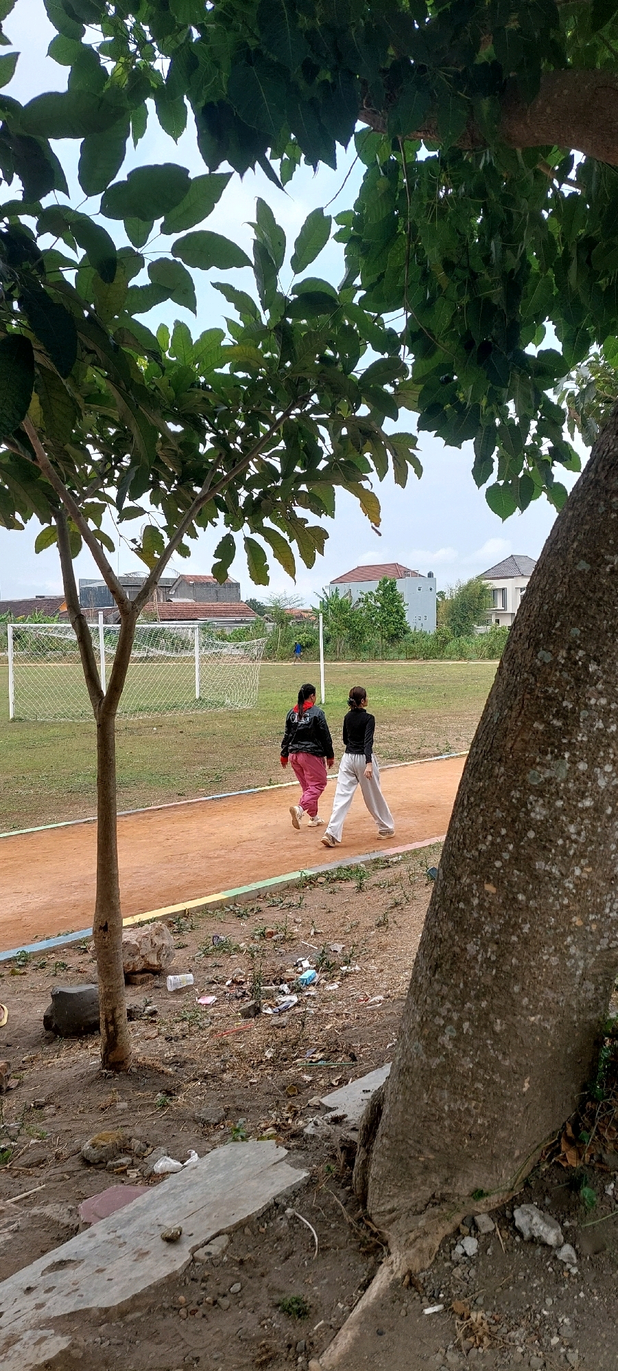 A vertical, medium shot captures an outdoor scene with two women walking on a dirt track that runs through a grassy field. The track is a reddish-brown color with a thin, multicolored border along its edge. The women, viewed from behind, are walking in the same direction on the track. The woman on the left wears a dark jacket with a red collar and bright pink pants. The woman on the right wears a black long-sleeved shirt and loose, light-colored pants.
Behind the track and the women is a green, grassy field with a white soccer goal visible in the background on the left. Further in the distance, several houses with tiled roofs are scattered among trees, indicating a residential area adjacent to the field. The sky is overcast.
In the foreground, on the left side of the image, a young tree with visible branches and green leaves partially obscures the view of the field. The ground in the immediate foreground is a mix of dirt, scattered rocks, debris, and a broken concrete slab. A large tree trunk, with rough bark and visible roots, dominates the right side of the frame, also partially obscuring the background. The overall scene suggests a recreational area within a semi-urban or suburban environment.