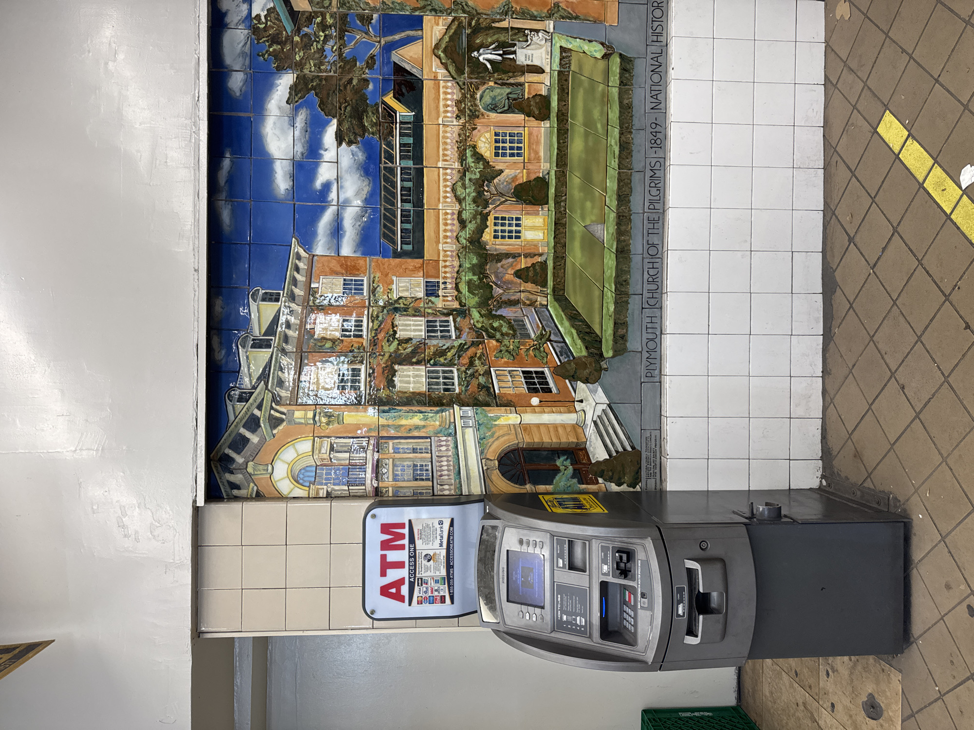 This image shows a portion of the subway station at Clark Street station in Brooklyn Heights, New York City. The station is decorated with tile mosaics. A large mosaic of the Plymouth Church of the Pilgrims, a National Historic Landmark, sits above an ATM. The ATM is gray and has a sign that reads "ATM ACCESS ONE." The ground is tiled.