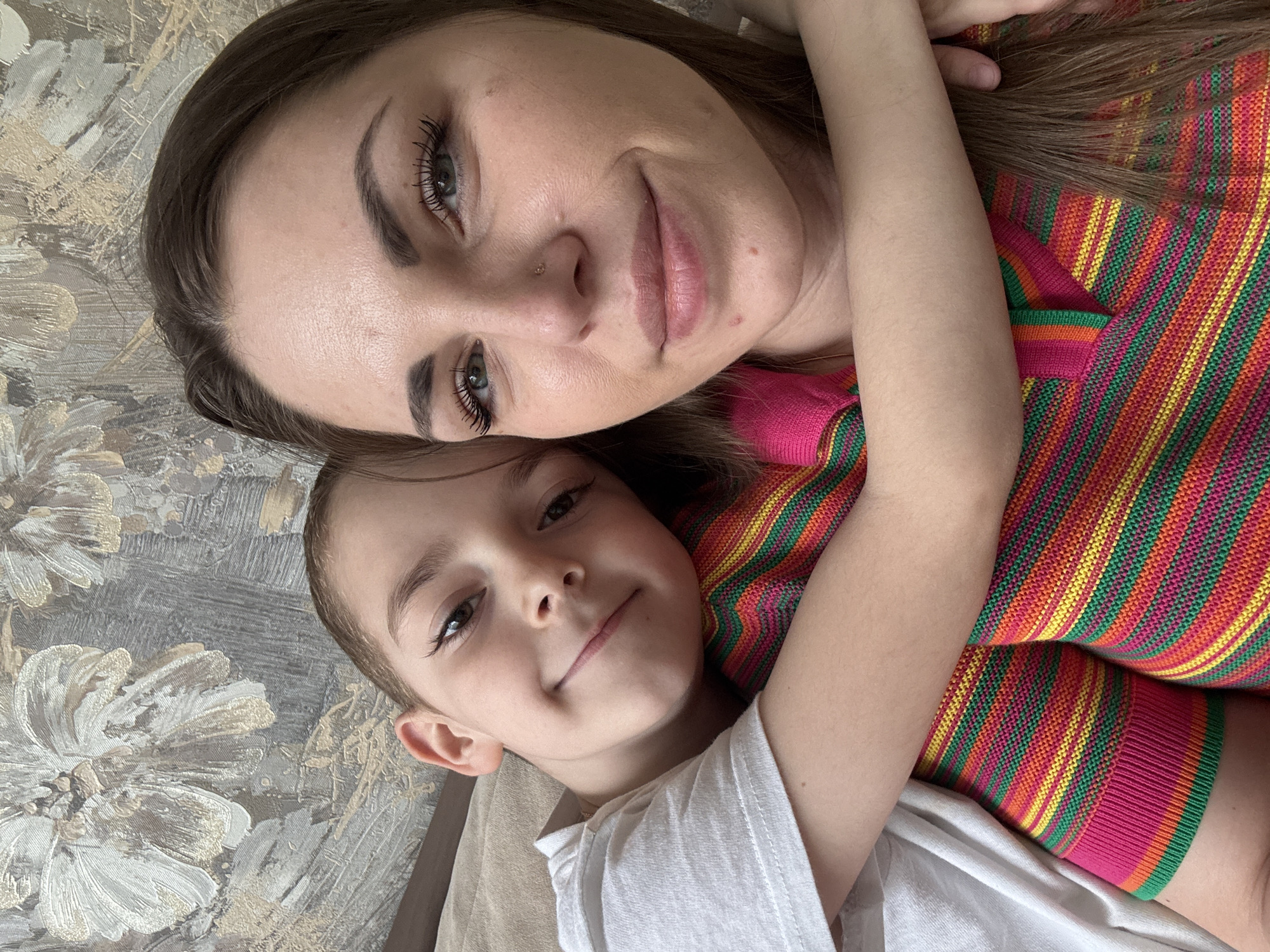 A woman and a child are lying down on a surface with a floral pattern, possibly a bed or sofa. The woman is at the top of the frame and is wearing a colorful striped shirt with pink, orange, yellow, and green stripes. She has dark hair, fair skin, and is smiling. The child, likely a young boy, is at the bottom of the frame wearing a white t-shirt. He is also smiling and appears to be embracing the woman. The background features a textured surface with a floral pattern, possibly wallpaper or fabric. The location is Раздолье, Russia.