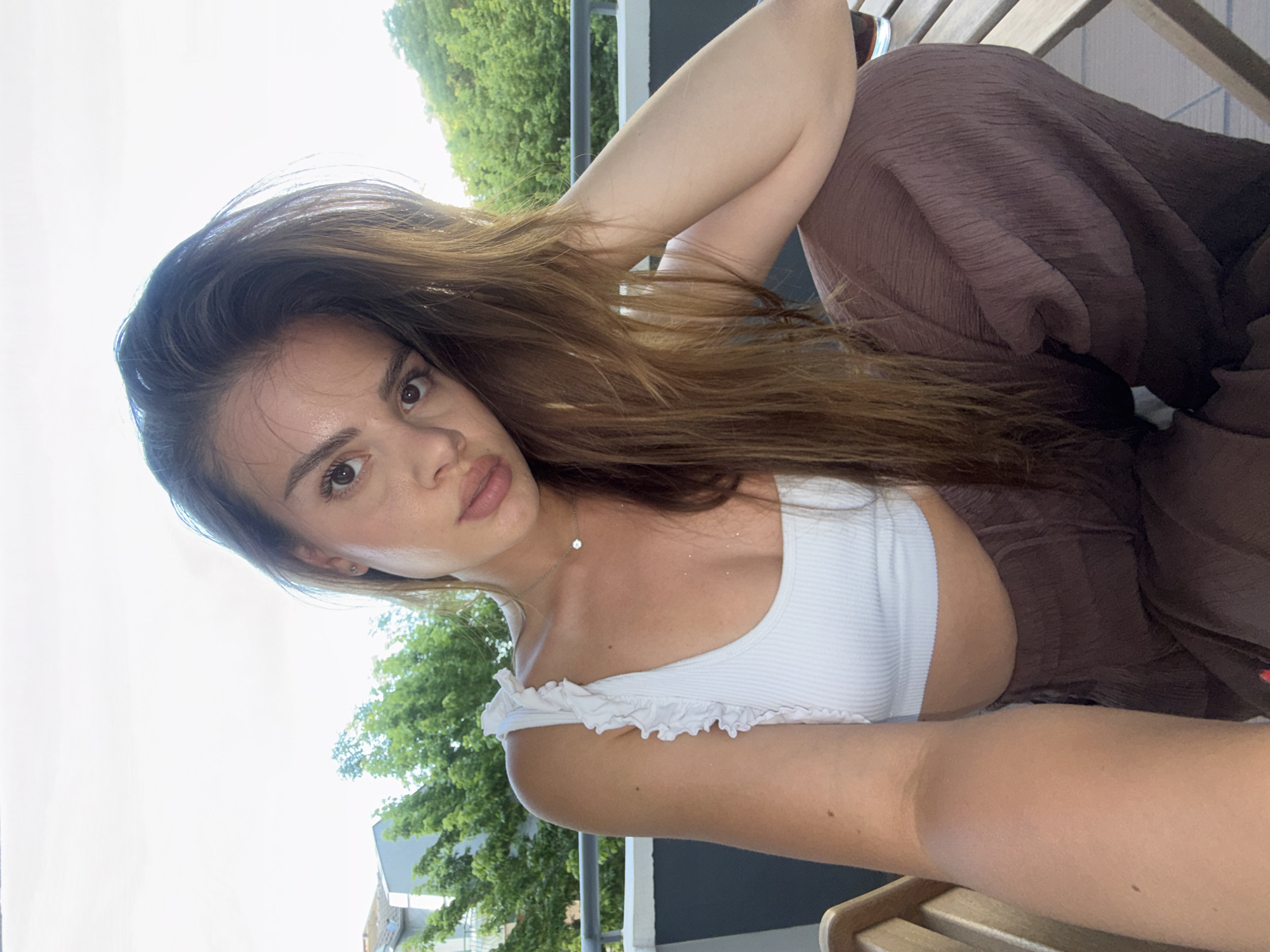 The image captures a young adult female posing for a direct-to-camera selfie on an outdoor balcony in Plovdiv, Bulgaria. She has long, dark brown hair styled to her right, and a fair complexion with a natural sheen. She is wearing a white, ribbed crop top with ruffled straps and dark brown, wide-leg trousers. A small silver necklace with a single clear stone is visible at her throat. Her left arm (viewer's right) is bent, resting on her raised knee, while her right arm (viewer's left) extends out of frame to hold the camera. The background features a dark metal balcony railing, followed by verdant green trees and the muted facades of distant buildings under a bright, overcast sky. Fragments of wooden slatted furniture are visible to her left and right, including a glass containing a dark liquid on the right. The lighting is natural and bright, suggesting daytime conditions.