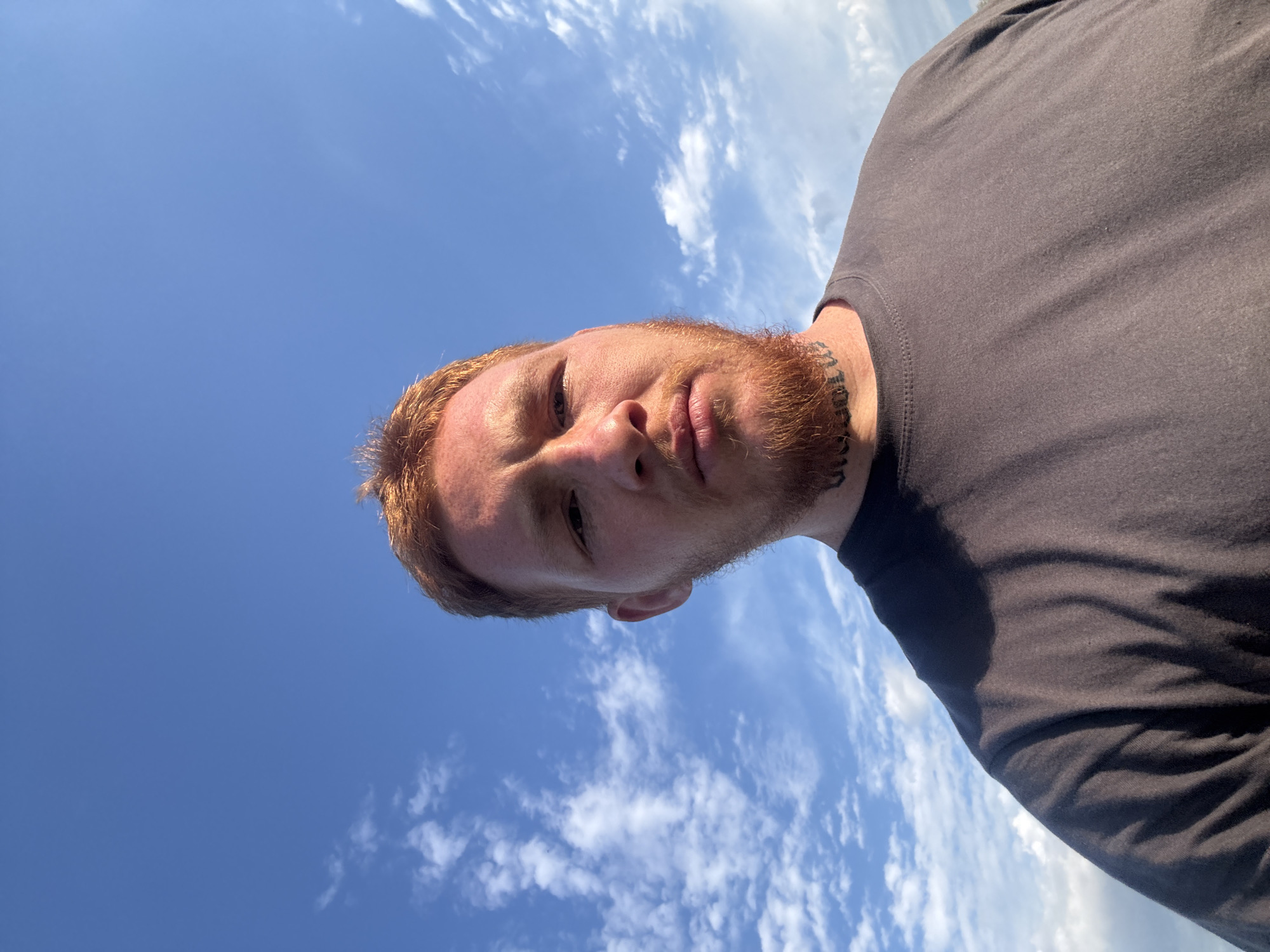 A selfie taken from a low angle shows a man with reddish-brown hair and a beard looking up at the camera against a bright blue sky with scattered white clouds. He is wearing a dark gray t-shirt. A tattoo is visible around his neck. The background suggests an outdoor setting with an open sky.