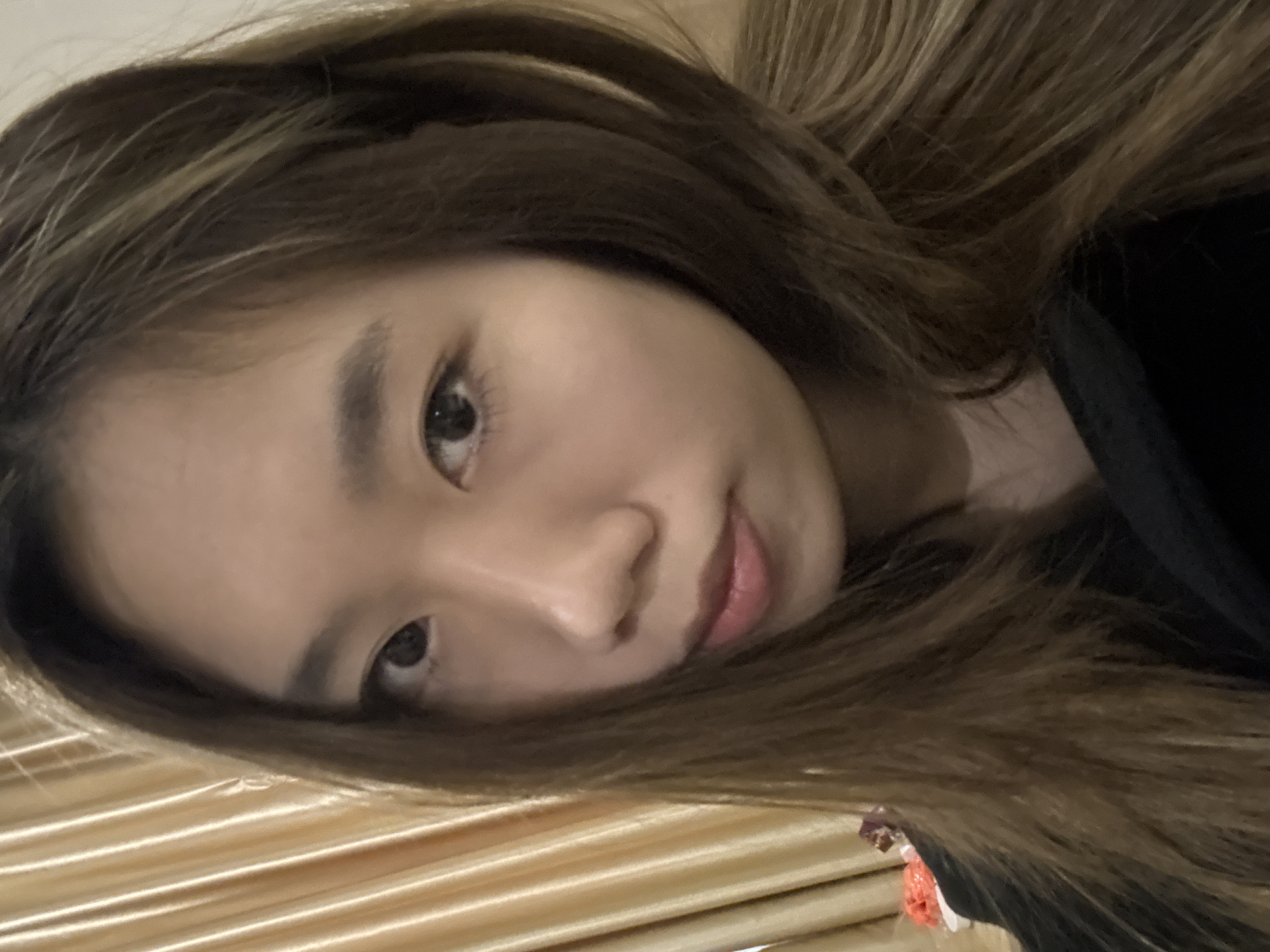 A young woman with fair skin and brown hair with lighter highlights looks directly at the camera. The lighting is soft and diffused. The background features a gold, textured surface, possibly a bedspread or draped fabric. The subject is partially obscured by her hair, and a hint of a dark-colored garment is visible at the right edge of the image. The setting cannot be determined from the image. There is no information related to Dunhuang City, China.