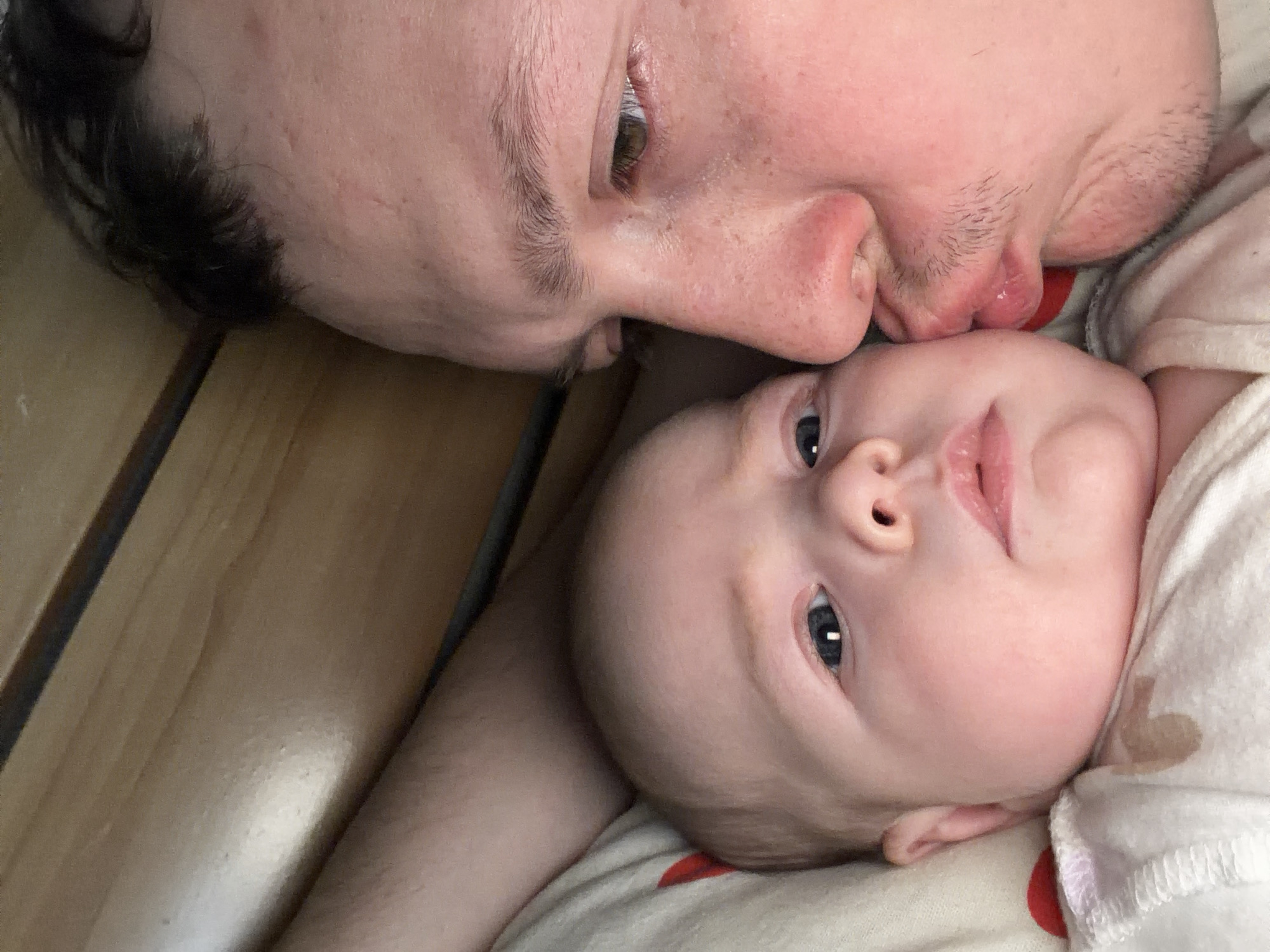 A close-up, tilted photograph shows a fair-skinned man and baby. The man, positioned above the baby, is in the process of kissing the baby's forehead/face. His eyes are open, looking slightly away from the camera, and his facial expression is gentle. He has dark hair with a receding hairline and some stubble. The baby's face is directed upwards, its eyes wide open and focused on something slightly above. The baby is wearing a light-colored garment with a pattern of hearts and possibly other shapes. The background includes what seems to be a wooden surface. The image does not provide details to confidently identify the location as the city of Chaikovsky, Russia. It is a domestic, intimate scene between a father and child.