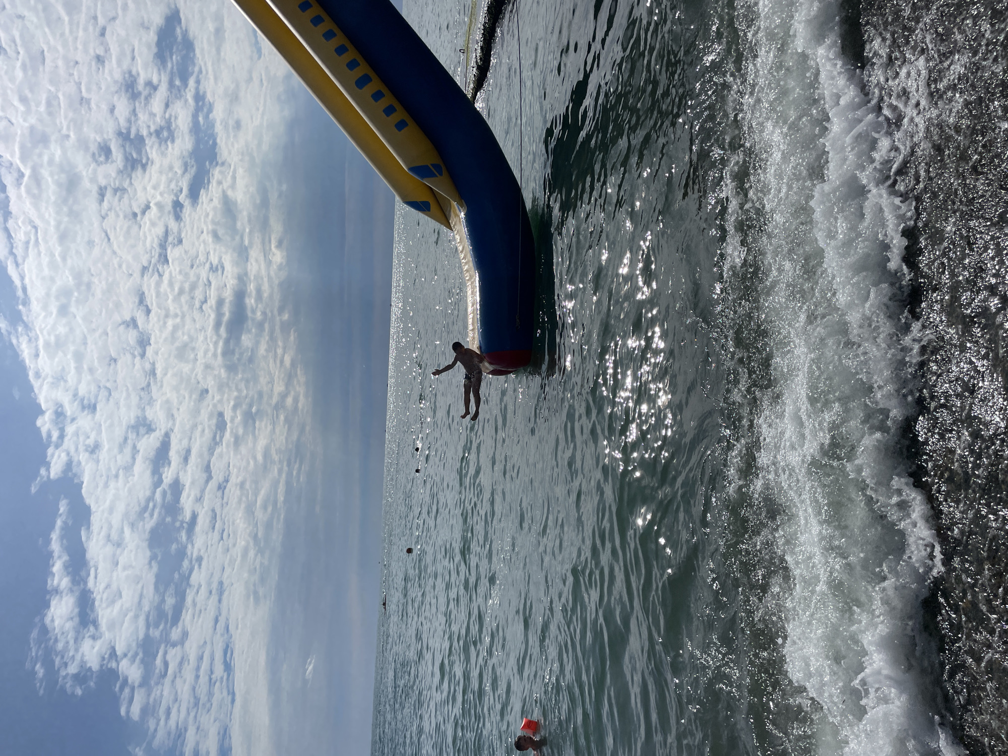 The media file depicts a sunny day at a beach in Sochi, Russia. The scene is dominated by the ocean, with waves crashing against what appears to be an inflatable structure. A person is seen jumping from the structure into the water, suggesting a recreational activity like water sliding or jumping. The sky is partly cloudy, with fluffy white clouds scattered across a light blue expanse. Sunlight glints off the water's surface, creating a shimmering effect. In the distance, other individuals can be seen in the water, some appearing to be swimming or enjoying the sea. The overall atmosphere appears to be one of fun and leisure on a warm summer day. There is no visible text in the image.