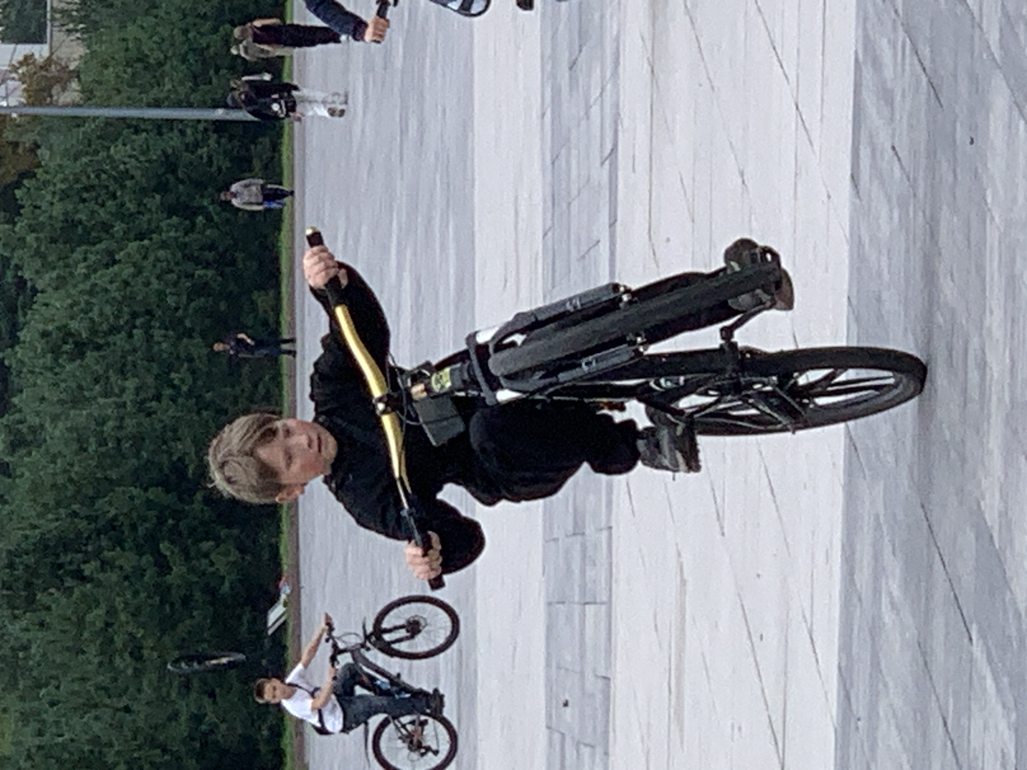A young boy with blond hair, wearing a black hooded jacket and black pants, performs a wheelie on a black bicycle with gold handlebars. His feet are on the pedals, and the front wheel of the bicycle is raised off the ground. In the background, another boy rides a bicycle, and several adults are walking in the distance. The scene appears to be an outdoor public space, possibly a plaza or park, paved with light-colored square tiles. Lush green trees form a dense backdrop behind the people and the open area. The lighting suggests it is daytime, possibly overcast. This image was taken in Prokopyevsk, Russia.