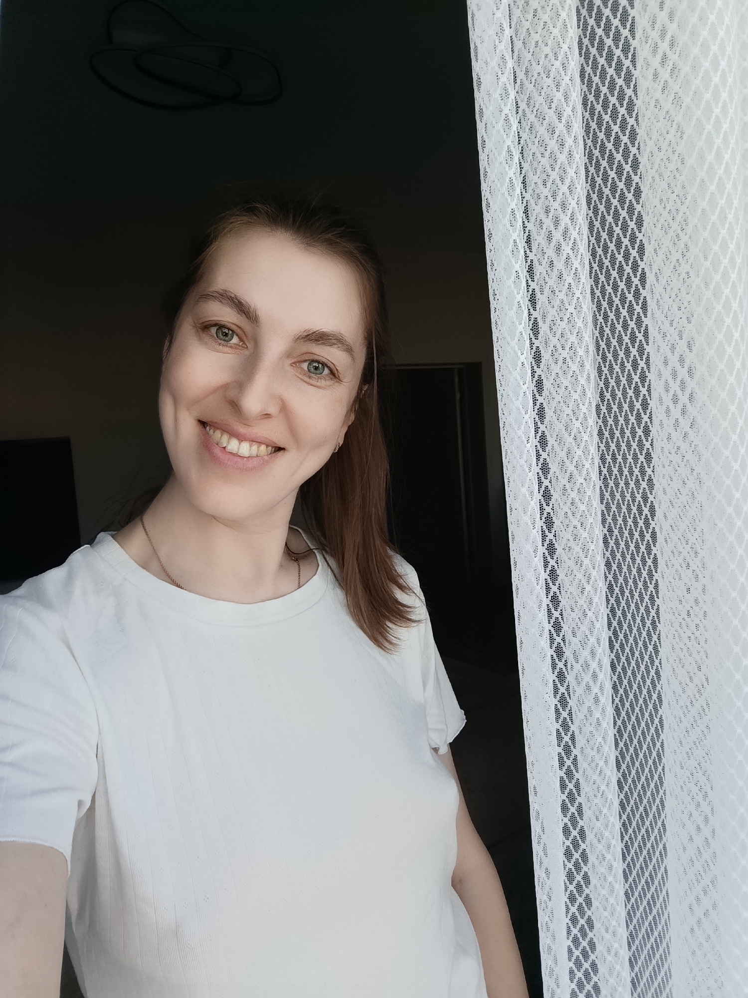 A Caucasian woman with fair skin, light brown hair, and green eyes is smiling into the camera. She is wearing a white t-shirt and a gold necklace. The woman appears to be standing indoors, next to a window covered with white net curtains. There's a dark doorway behind her. The location may be in the city of Chaikovsky, Russia, as requested.