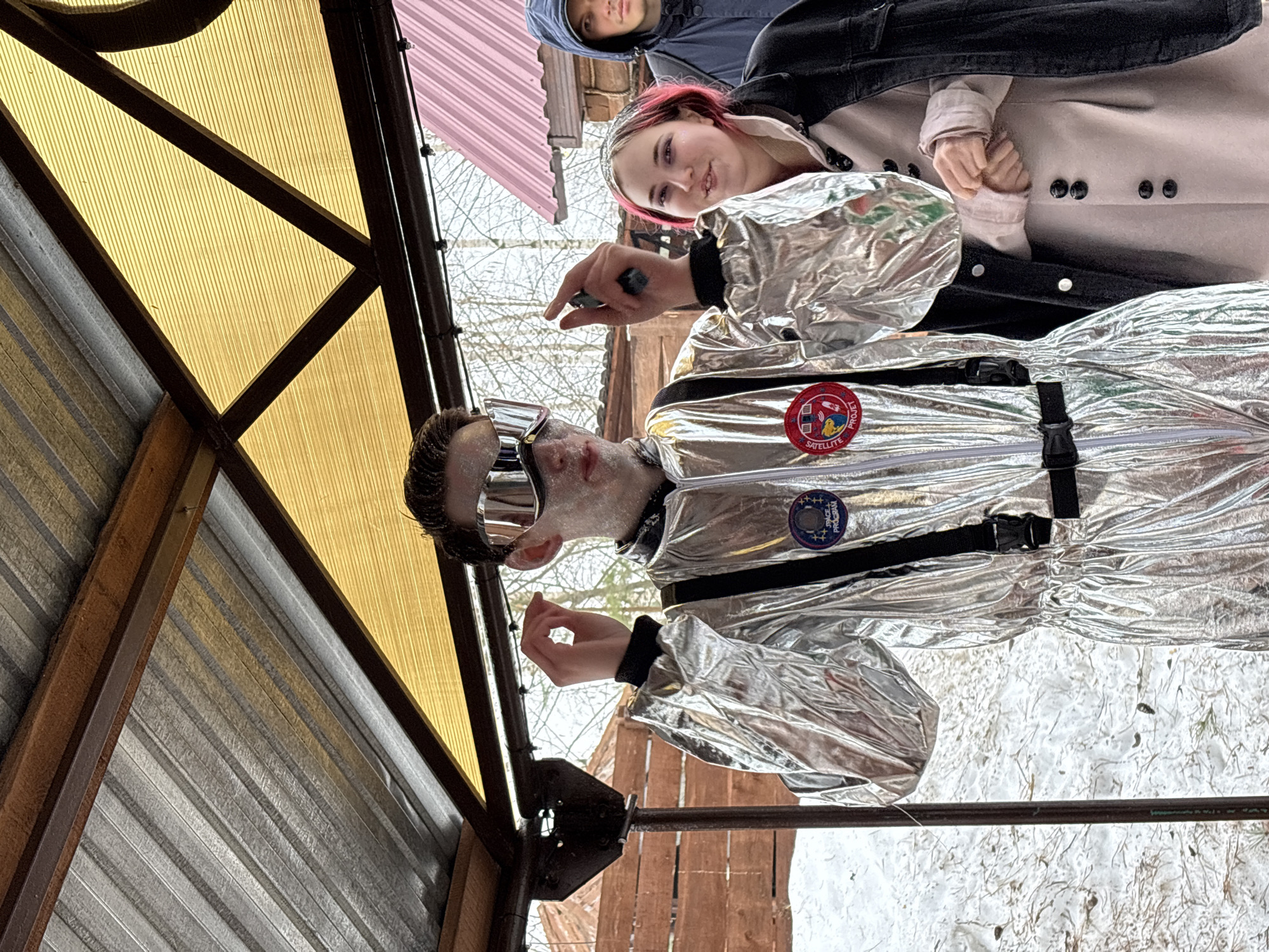 The image shows three people outdoors in what appears to be a park or wooded area in Chaikovsky, Russia. One person is dressed in a shiny silver spacesuit-like costume, complete with a buckle strap across the chest and patches indicating "Satellite Project" and "Space Program." They are wearing silver goggles and have some kind of white substance on their face. This person is holding something small between their fingers. Behind the costumed individual is a girl with pink-streaked hair, wearing a light-colored coat with black buttons. Behind her is a person in a hooded jacket. The background shows bare trees and snow-covered ground. The scene is partially framed by the structure of a gazebo with a translucent yellow roof.