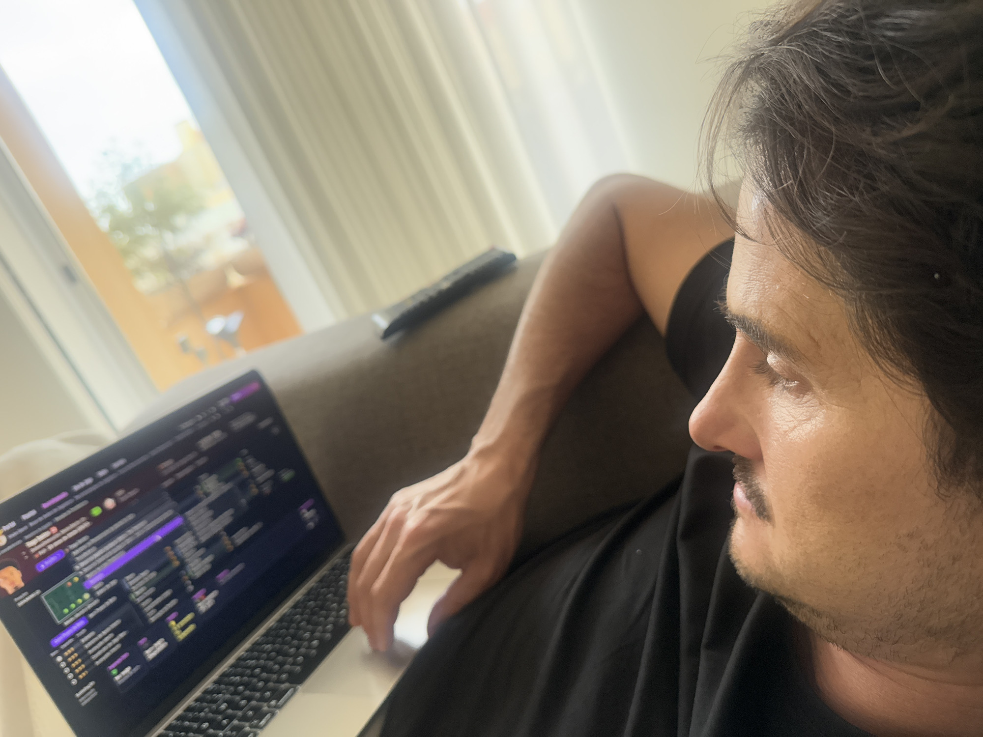 A man with dark hair and a mustache is reclined on a light-colored couch, looking at a laptop screen. The laptop is positioned in front of him on the couch. The screen displays a website with purple and green colors. The man's right hand is near the laptop's trackpad. A black remote control rests on the couch behind the laptop. A window with white curtains is visible in the background. The exact location in the city of Vila Nova de Gaia, Portugal, cannot be determined from this image alone.