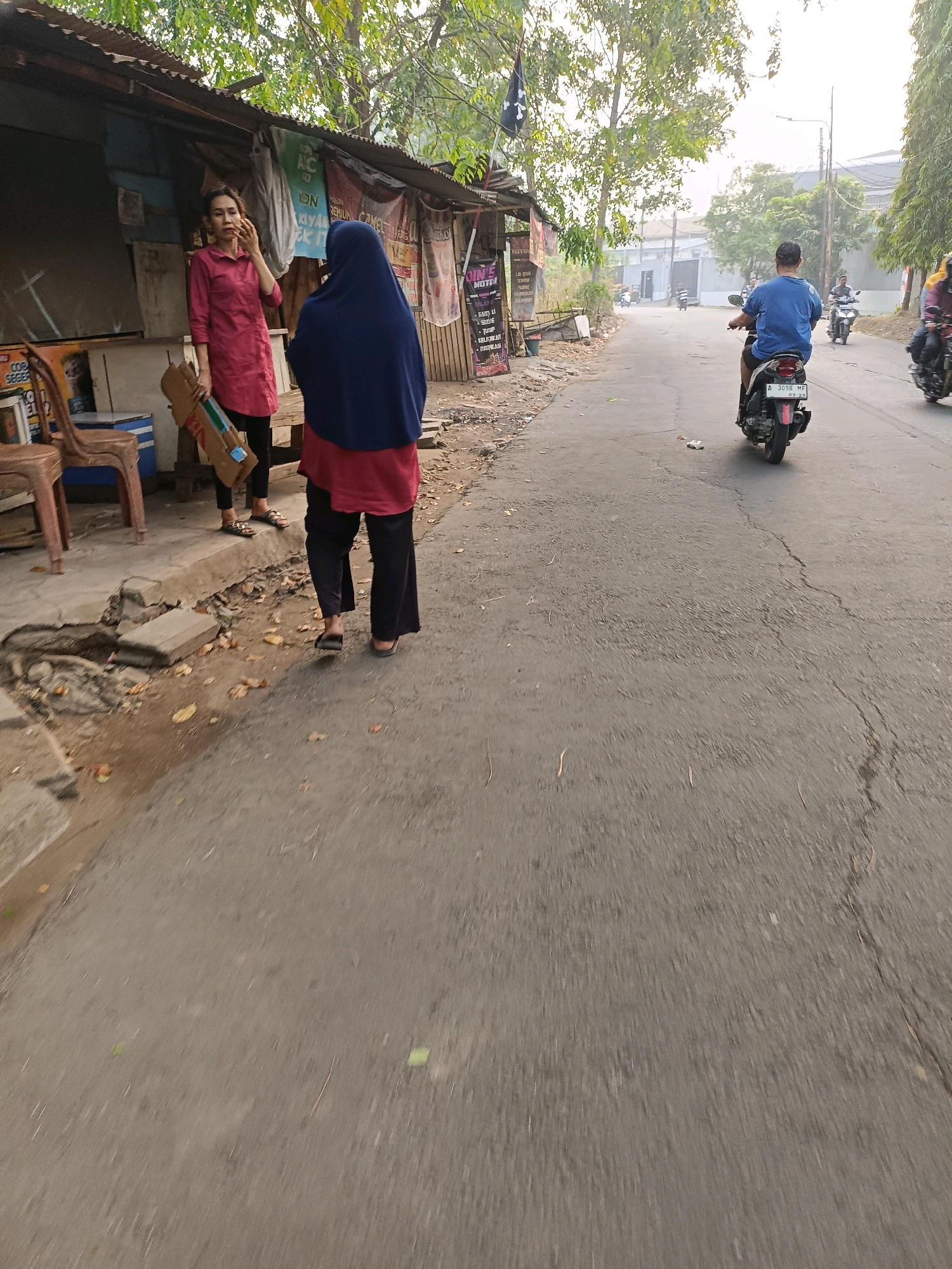 A person wearing a blue hijab and a pink shirt stands with their back to the camera on a street in Tangerang, Indonesia. The person is walking on a cracked asphalt road. To the left of the road, there is a small wooden structure with a tin roof, possibly a stall. Another person in a pink top is standing to the left of the stall, holding a cardboard tube. In the background, several people are riding motorcycles on the road, and there are trees and buildings. The sky is overcast.
