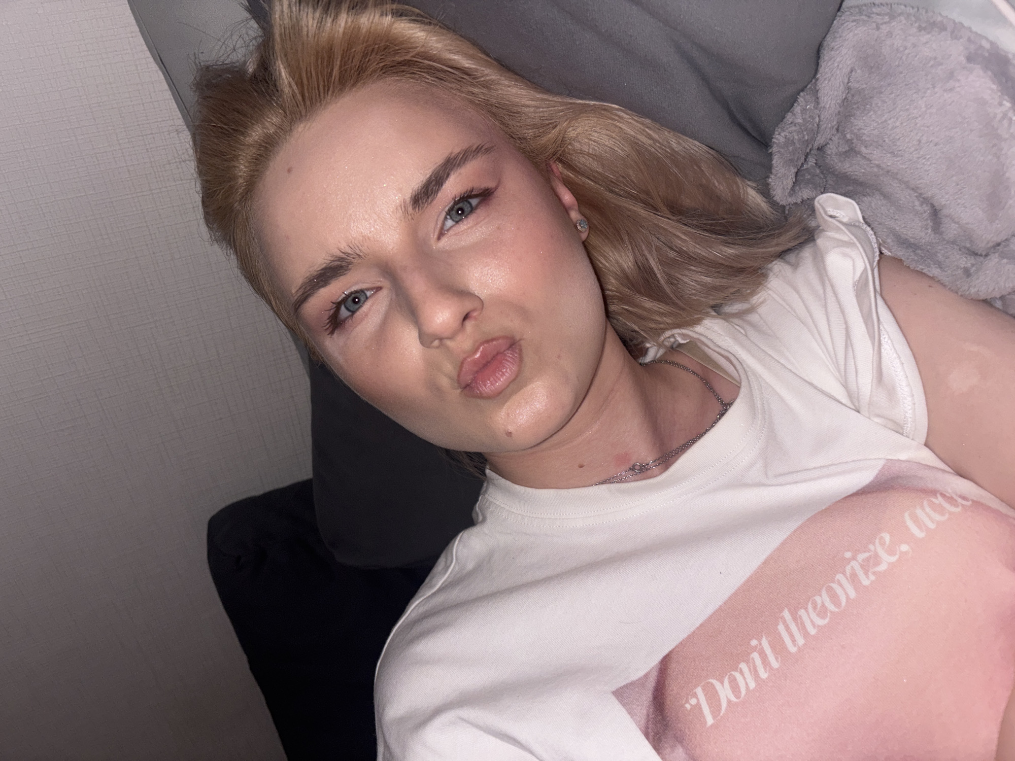 A young blonde female individual is depicted lying on her side in an indoor setting, likely on a couch or bed, in the city of Unknown, Russia. She is facing the viewer with her head slightly tilted, making a puckering or "kiss" face with her lips. Her eyes are open, revealing blue irises, and she has visible eye makeup, including eyeliner and light pink eyeshadow, with a slight shimmer on her eyelids and cheekbones. Her blonde hair is styled casually around her face and head. She is wearing a white short-sleeved t-shirt over what appears to be a light pink garment or a part of the shirt's design. White text on the pink area reads, "Don't theorize, act." A thin silver chain necklace is visible around her neck, along with a small red mark near her collarbone. A small stud earring is present in her right ear. The background features a textured gray wall behind her, and she is resting on dark gray or black cushions/furniture. A light gray, plush blanket is partially visible to her right. The lighting suggests an indoor, possibly evening, environment.