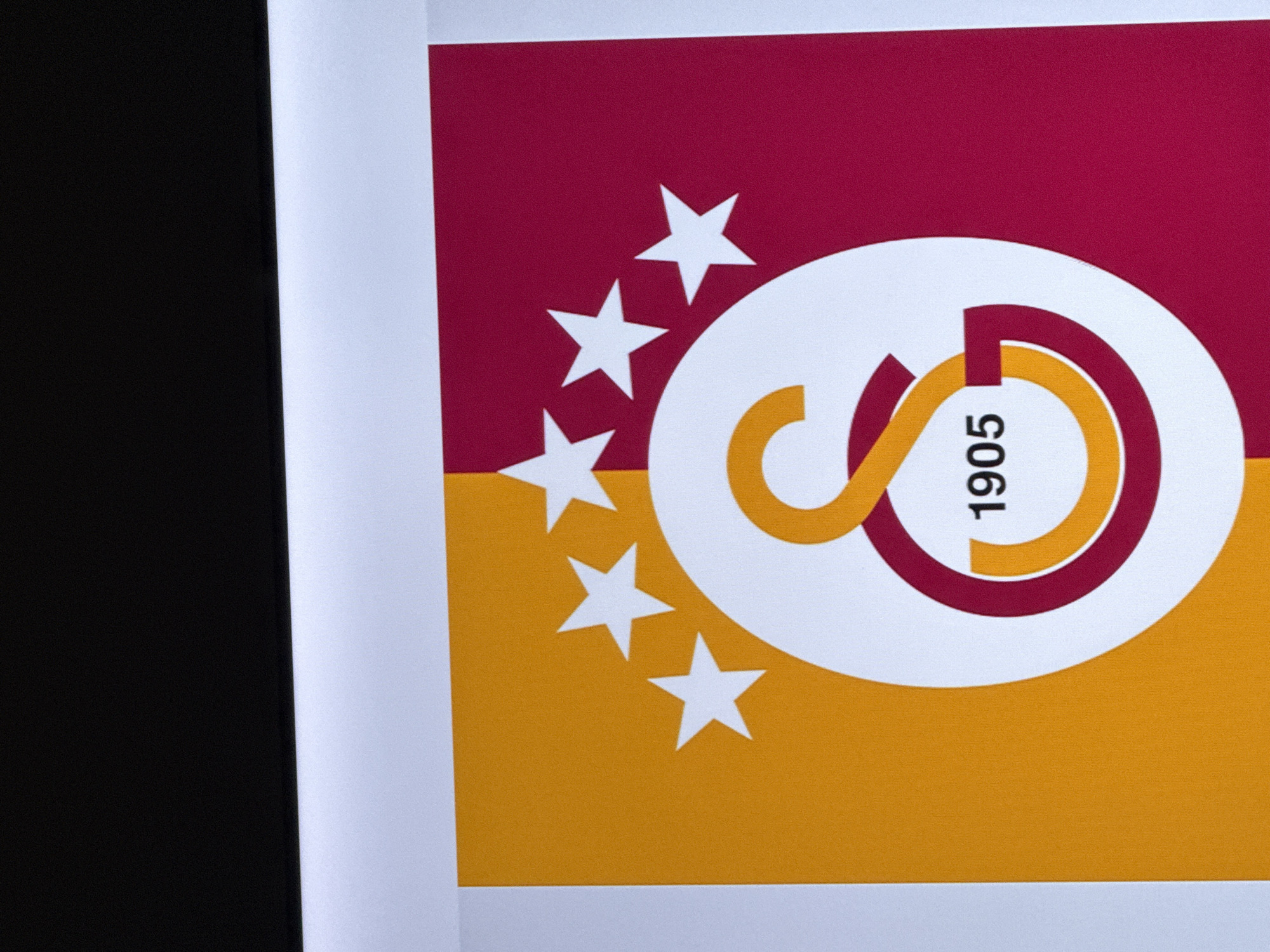 A close-up shot shows a Galatasaray S.K. flag divided horizontally into claret red (top) and yellow (bottom). A white circle in the center features intertwined red and yellow shapes, resembling "GS" with "1905" vertically positioned. Five white stars are positioned on the left, partially overlapping the circle and the background colors. The left edge of the image contains a black area next to a white border.