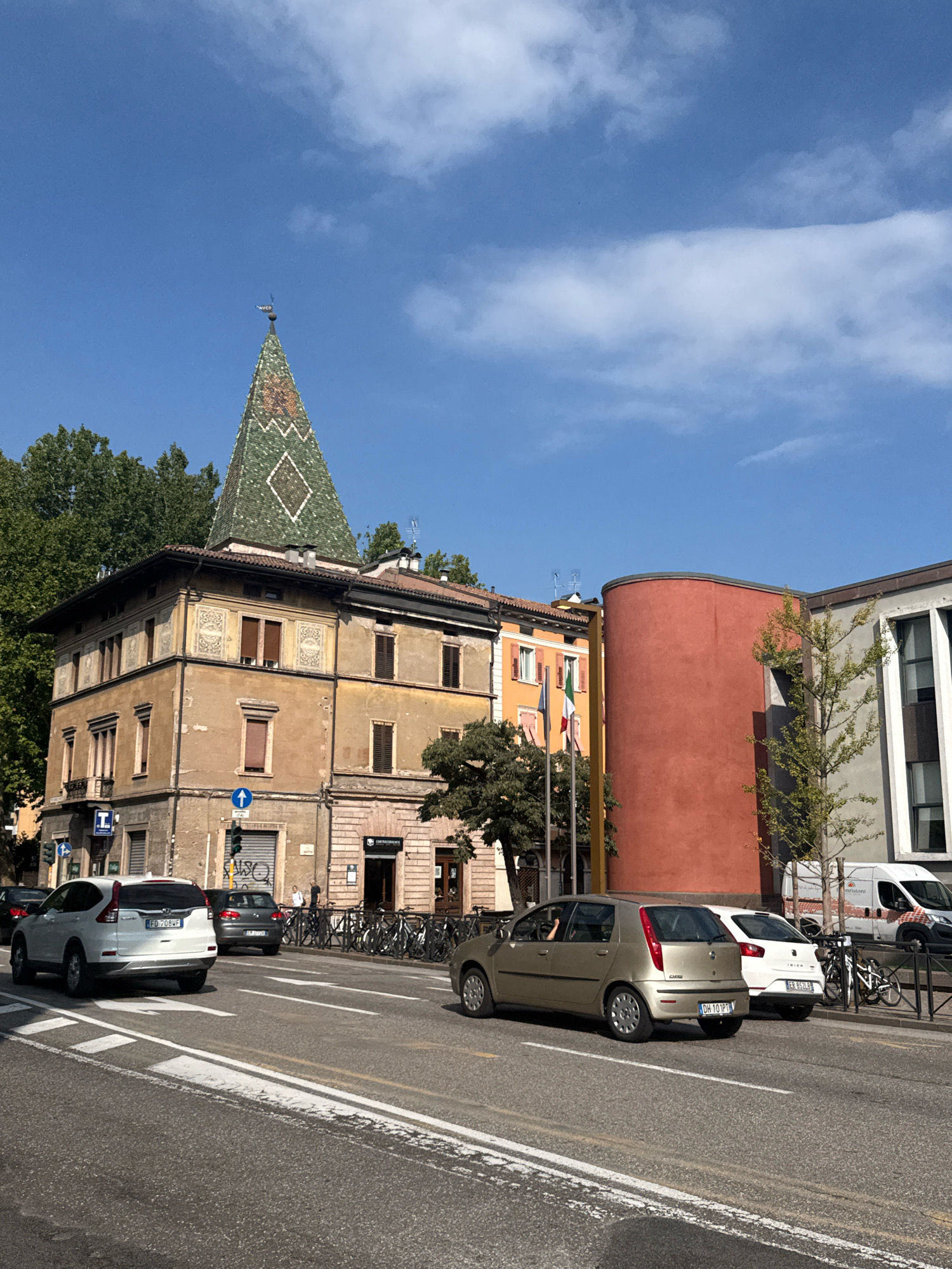 In Trento, Italy, a view down a street shows several cars on the road and parked. A white SUV, a dark gray hatchback, and a light gold hatchback are driving. Parked cars, bicycles, and a white van are also visible. Buildings line the street, one with a distinctive green, pointed roof and ornate decoration. Another building has a curved, red facade. Italian and other flags are displayed. The sky is blue with scattered clouds. Trees with green foliage are present.