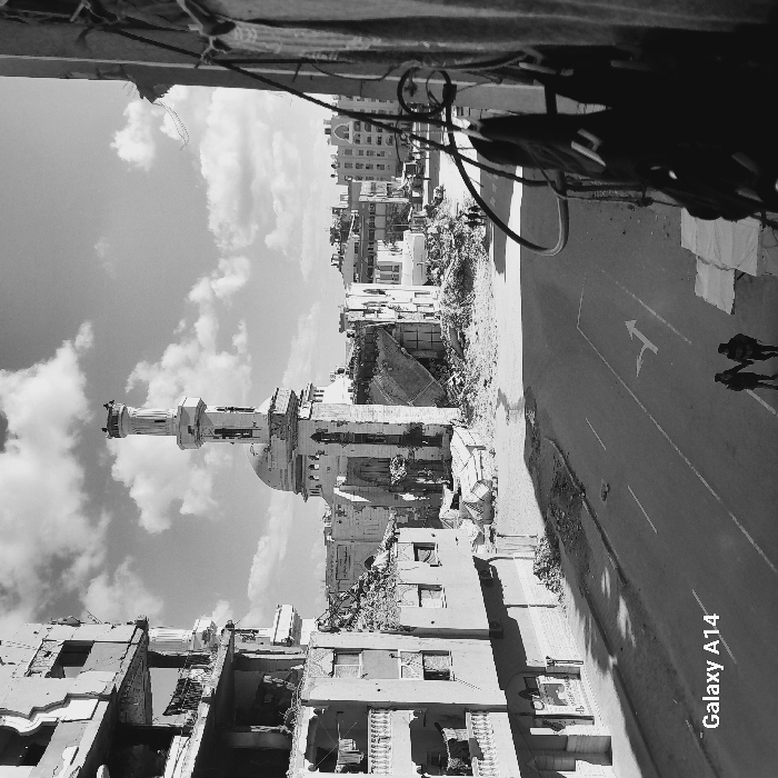 This black and white image depicts a scene of extensive urban destruction in Khan Yunis, Palestinian Territory, captured from an elevated perspective. The focal point is a severely damaged mosque in the middle ground, featuring a partially standing minaret with an intact top section and a large, partially collapsed dome. Its main structure appears gutted, revealing skeletal remains and significant debris.
To the left, a multi-story building exhibits extensive structural damage, with walls and floors blown out, exposing internal elements. Clothes are visibly hanging on a balcony railing on one of its lower floors. The ground surrounding the mosque and adjacent buildings is covered in large piles of rubble and debris.
An asphalt street runs through the lower and middle sections of the image, showing a white painted arrow indicating a turn. Two individuals, holding hands, walk on the street in the lower left foreground, moving away from the viewer. Further down the street, near the mosque, a group of three additional individuals is visible. In the immediate foreground on the right, structural elements of another damaged building are visible, with exposed wires or cables hanging. The background shows additional urban structures, some appearing intact, others partially damaged, under a partly cloudy sky. A "Galaxy A14" watermark is present in the bottom left corner.