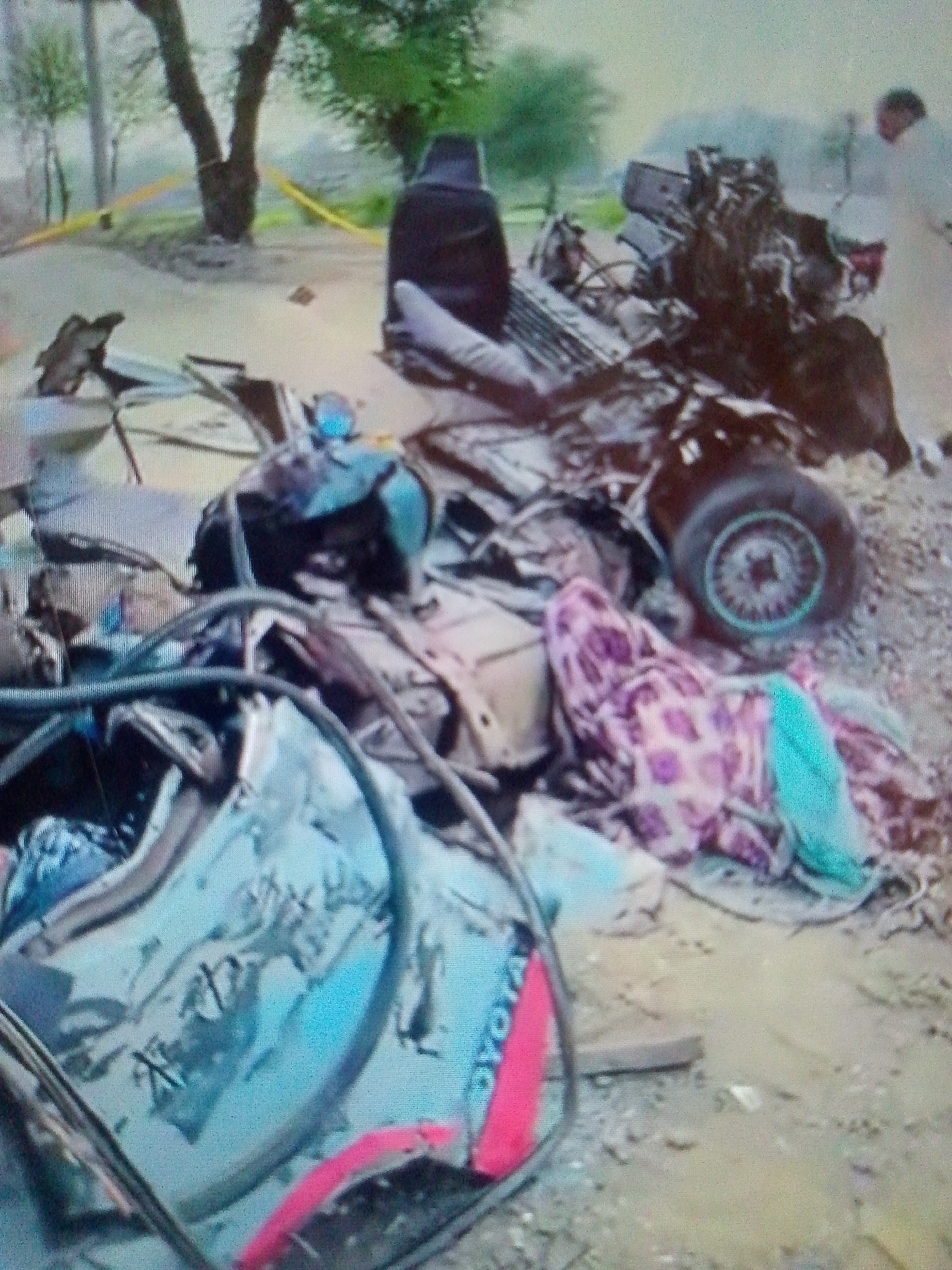 The image depicts the aftermath of a severe car accident in an indeterminate location. The wreckage of a vehicle, identified by "TOYOTA" lettering on a fragment, is scattered across the ground. Visible are parts of the car, including a wheel, a seat, and twisted metal. A patterned cloth lies amidst the debris. A person in a white garment stands in the background, observing the scene. Yellow tape is visible, potentially marking the area as a crime scene. The surroundings include trees and what appears to be a roadside environment.