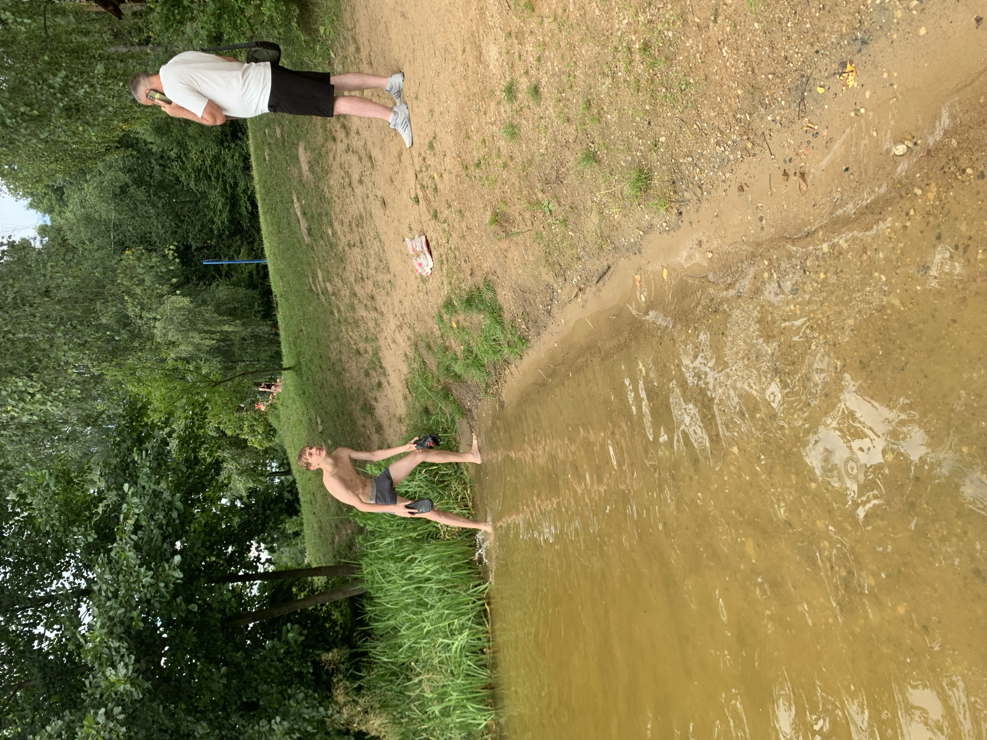 The image depicts a scene by a body of water, likely a lake or river, in a natural setting. Two individuals are visible. In the foreground, a shirtless young person wearing dark shorts stands at the edge of the water, with their feet touching the muddy shore and the water. They are looking towards the viewer and appear to be posing or about to enter the water. Behind them, to the left, is a dense area of green trees.
Further in the background, a man wearing a white t-shirt and black shorts is standing on a sandy bank, holding a phone to his ear. He is looking down and appears to be engaged in a conversation. His shoes, a pair of white sneakers with red accents, are placed on the ground near him. The surrounding area includes sandy soil with patches of grass and scattered pebbles. The overall impression is of a casual outdoor activity during daylight hours. The sky is not clearly visible, but the lighting suggests it is daytime. The location is identified as Грозаўскі сельскі Савет, Belarus. No visible text is present in the image.
