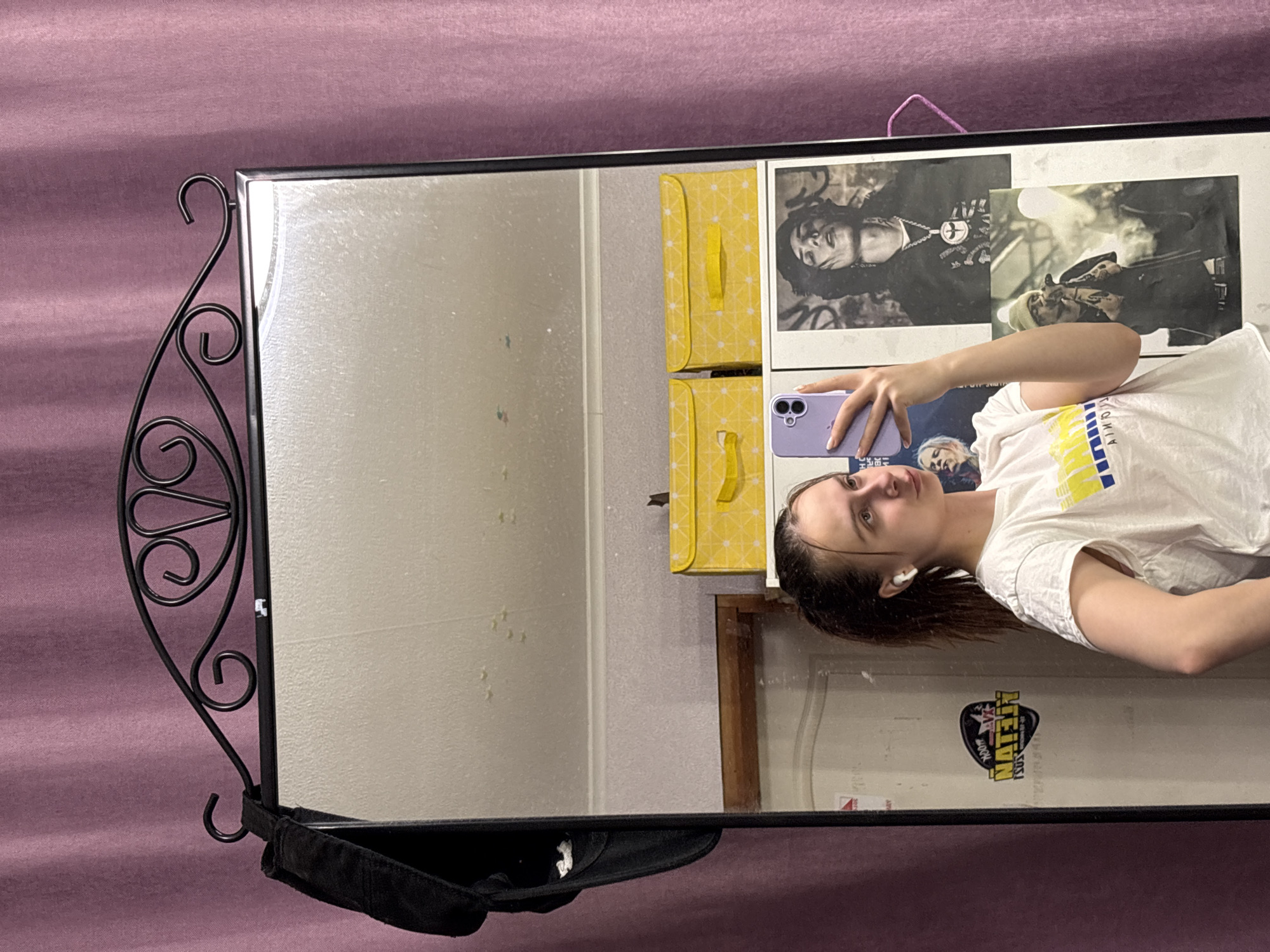 A young woman is taking a selfie in a mirror. The mirror has a black metal frame with decorative elements. A black baseball cap is visible at the bottom of the frame. The background includes a mauve-colored wall, yellow storage boxes, black and white portraits on the wall, and a wooden shelf with a sticker. The scene is likely indoors. There is no concrete evidence that the location is Прикамский, Russia.