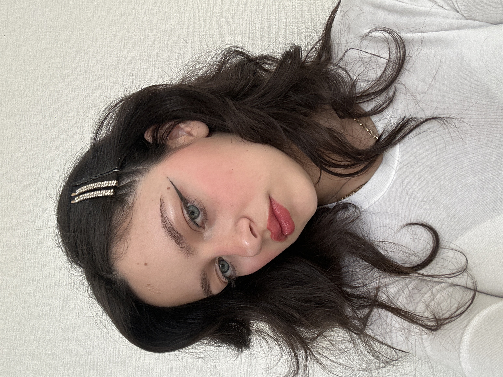 A young woman with dark brown hair, fair skin, and light eyes is depicted in this portrait-style image. She has a prominent winged eyeliner, rosy cheeks, and red lipstick. A bejeweled hair clip adorns her hair near her forehead. She is wearing a white top, and a gold necklace is visible around her neck. The background is a plain, light-colored wall. The image appears to be taken indoors. The city of Yerevan, Armenia, is not discernible from the provided visual information.