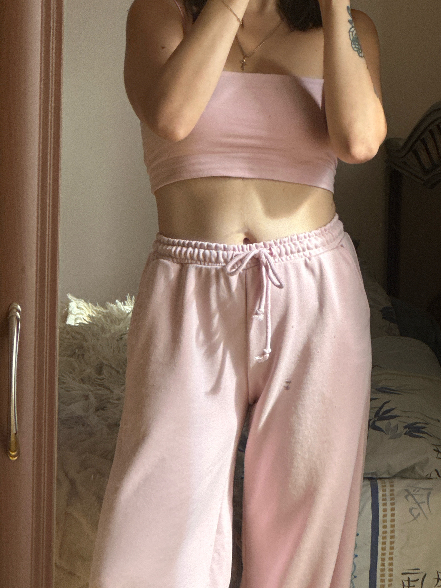 The image displays a medium shot of a person wearing a pink strapless top and pink sweatpants. The person is standing in what appears to be a bedroom, with a bed visible in the background. The lighting suggests it is daytime, with sunlight casting shadows. There is a dresser or wardrobe to the left of the frame, and a fluffy white rug in front of it. The person's hands are raised, obscuring their face, and a tattoo is visible on their arm. The overall scene is casual and domestic. There is no identifiable information in the image that would allow for the location to be determined as Gyumri, Armenia.