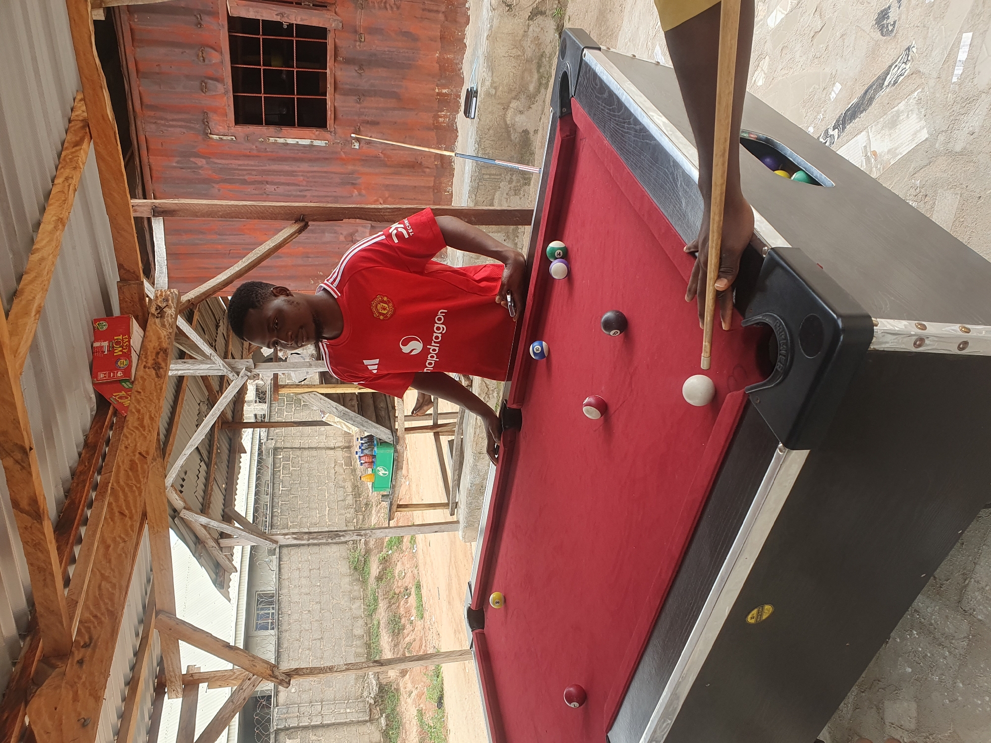 In Lokoja, Nigeria, under a rustic, open-air shelter with a corrugated metal roof supported by wooden beams, a young Black man wearing a red Manchester United jersey is playing pool. He is positioned behind the pool table, holding a cue stick and looking intently at the red felt playing surface, which has several billiard balls scattered across it. The environment appears to be an outdoor recreational area, with a weathered corrugated metal structure visible in the background, a brick wall, and sandy ground. The lighting suggests it is daytime.