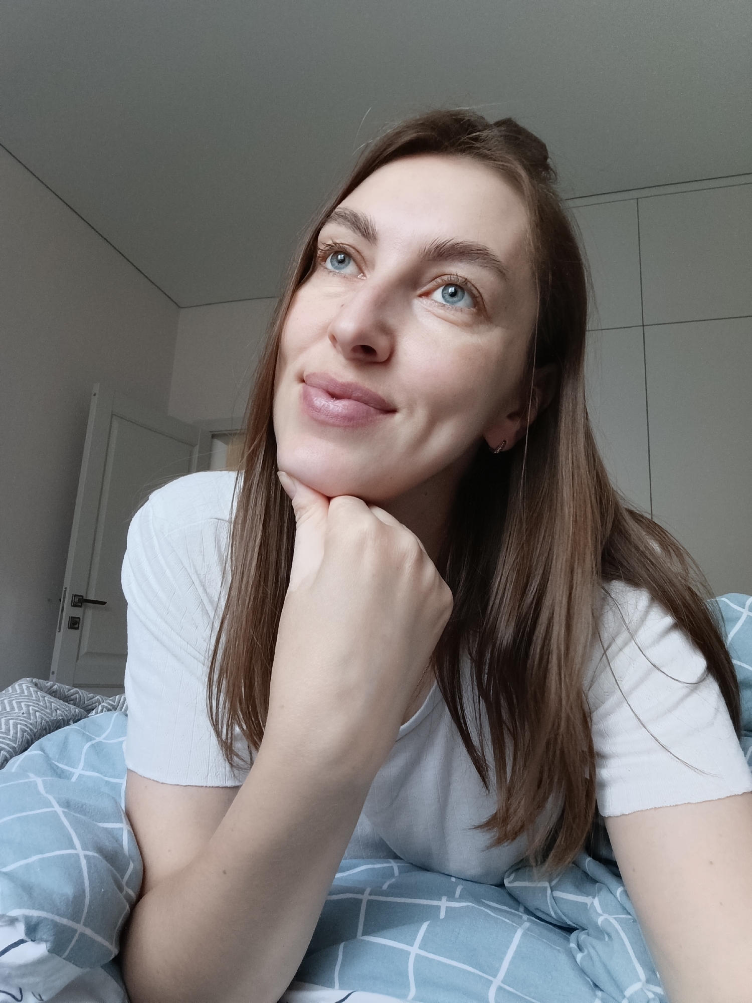 A woman with light skin, blue eyes, and light brown hair is indoors, resting her chin on her right hand. She is wearing a white t-shirt and is lying on a bed with blue and white patterned bedding. The room has a white door in the background on the left side and white cabinets on the right. While it is not possible to confirm the location as Chaikovsky, Russia, from the image, the setting appears to be a residential bedroom.