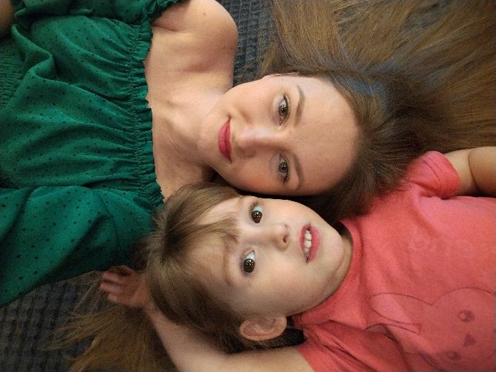 An overhead photograph captures an intimate portrait of an adult and a young child, both lying down with their heads close together and looking up towards the camera. The subjects are positioned diagonally across the frame, against a dark, textured background that appears to be a carpet or rug.
The adult, on the left side of the image, has light skin, striking light brown or hazel eyes, and a gentle smile accentuated by red lipstick. Her long, medium brown hair is spread out behind her head. She is wearing a dark green, off-the-shoulder top that features small black polka dots and a smocked, elasticated neckline and sleeves.
Nestled close to the adult, on the right side of the frame, is a young child. The child also has light skin and distinct brown eyes that are wide open, conveying a sense of curiosity or slight surprise. Her mouth is slightly open, revealing some teeth. She wears a plain, short-sleeved pink or coral t-shirt. Her lighter brown hair is also fanned out around her head, intertwining slightly with the adult's hair.
The composition suggests a tender and affectionate moment, likely between a parent and child, captured from a unique top-down perspective. The lighting is soft and even, highlighting their faces. No discernible text is visible in the image.