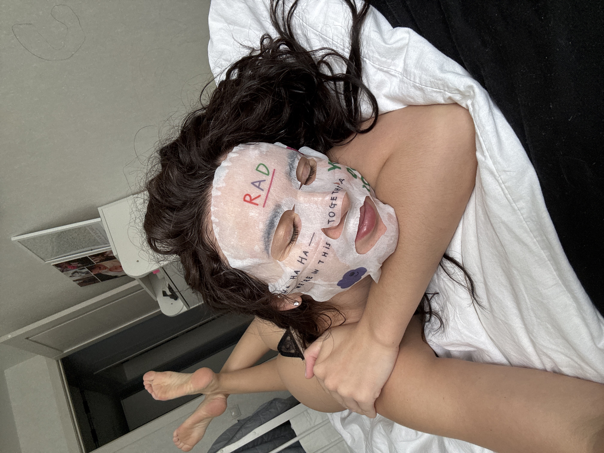 A person is depicted lying prone on a bed in an indoor setting. The subject's face is covered by a white sheet mask featuring colorful text and graphics, including "RAD" in red on the forehead, "HA HA — WE'RE IN THIS" below the right eye, "TOGETHER" below the left eye, and a purple cloud graphic on the left cheek. The subject's eyes are closed, and the mouth is slightly agape beneath the mask. Dark, wavy hair is spread around the subject's head. The subject wears black lace undergarments, specifically briefs, partially visible on the left hip. The left arm is bent, with the hand resting near the torso. The legs are bent at the knees, with the soles of the feet visible in the upper left of the frame. The bed is covered with white linen. The background shows a light-colored wall, a partially open white door, and a white shelf unit displaying multiple framed pictures. The specific location within the city of Chaikovsky, Russia, cannot be determined from the visual evidence.