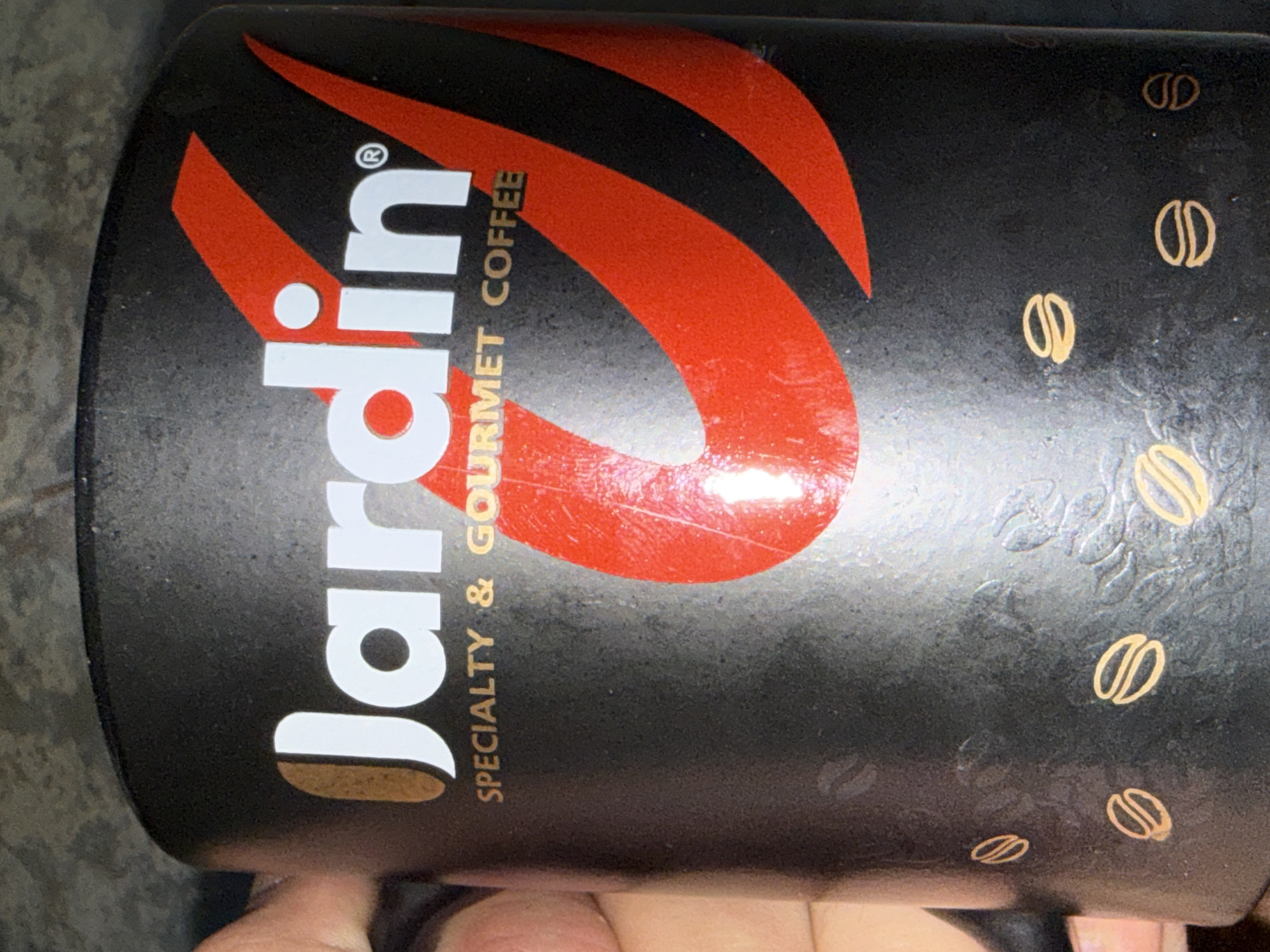 The image is a close-up, eye-level shot of a black cylindrical container, likely for coffee. The prominent branding features the word "Jardin" in large, white, sans-serif letters with a golden accent on the "J". Below this, the text "SPECIALTY & GOURMET COFFEE" is printed in smaller, golden, sans-serif letters. A stylized red and black graphic design, resembling curved stripes or flames, is visible on the right side of the container. Scattered across the lower portion of the container are small, golden illustrations of coffee beans. The background is a textured, dark surface that appears to be a tabletop or counter. The overall lighting is dim, with some reflections on the container's surface. There is no indication of the city of Cahul, Moldova, in the image.