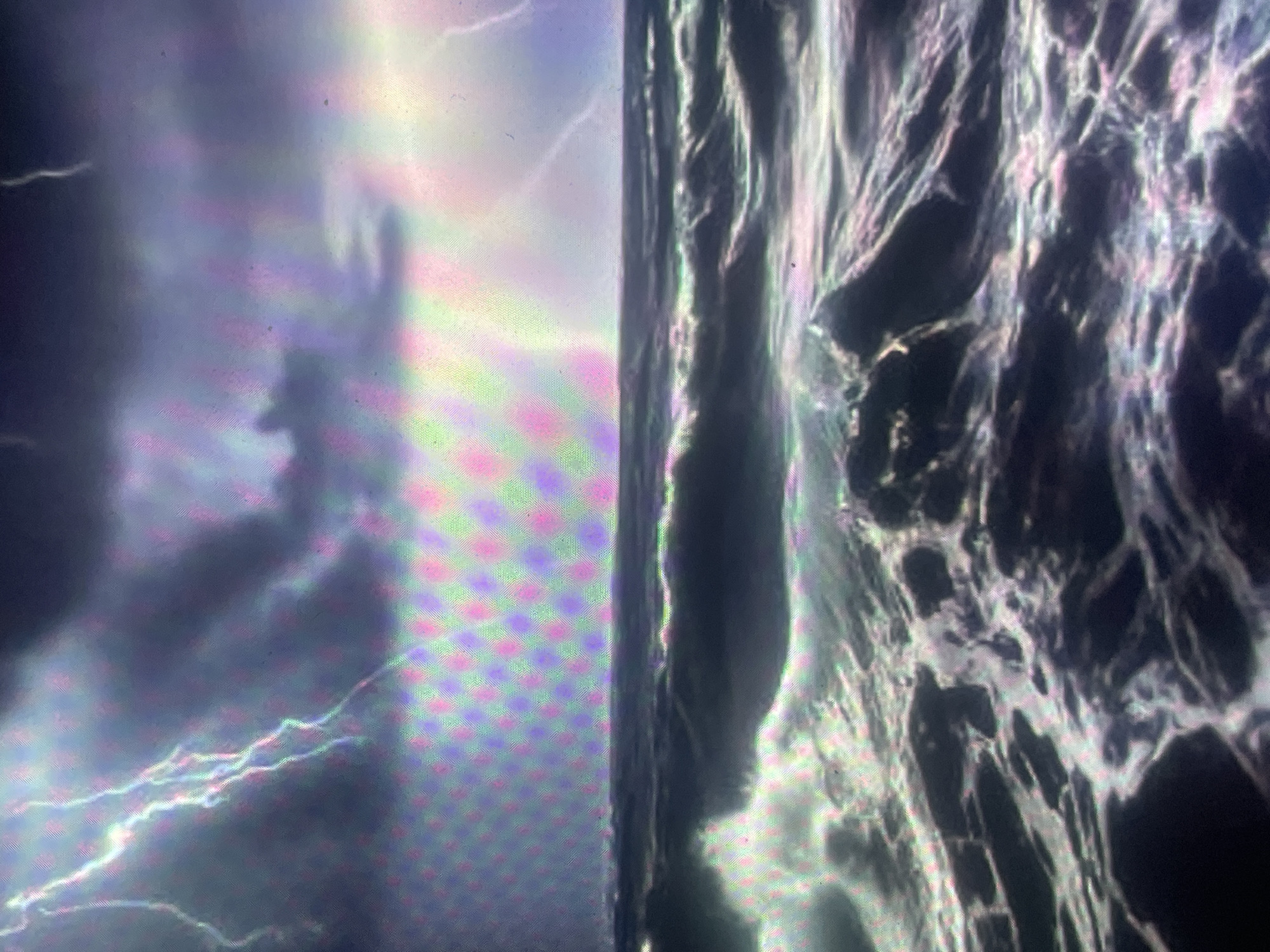This image is a photograph of a digital screen displaying a stormy open ocean scene. The sky is dominated by dark, heavy clouds, with multiple bolts of lightning striking across the upper left quadrant and a luminous glow on the right horizon. The sea below is rough, characterized by large, dark waves and white crests of foam, particularly prominent in the foreground. A distinct, repeating grid pattern of multi-colored subpixels (magenta, green, blue, and cyan) is visible across the entire image, a characteristic artifact of photographing a digital display. The depicted environment is an open ocean under severe weather conditions, devoid of any human presence, specific man-made objects, or interactions. In the context of Zelenyi Hai, Ukraine, this image is an observation of a digital screen presenting this severe weather landscape.