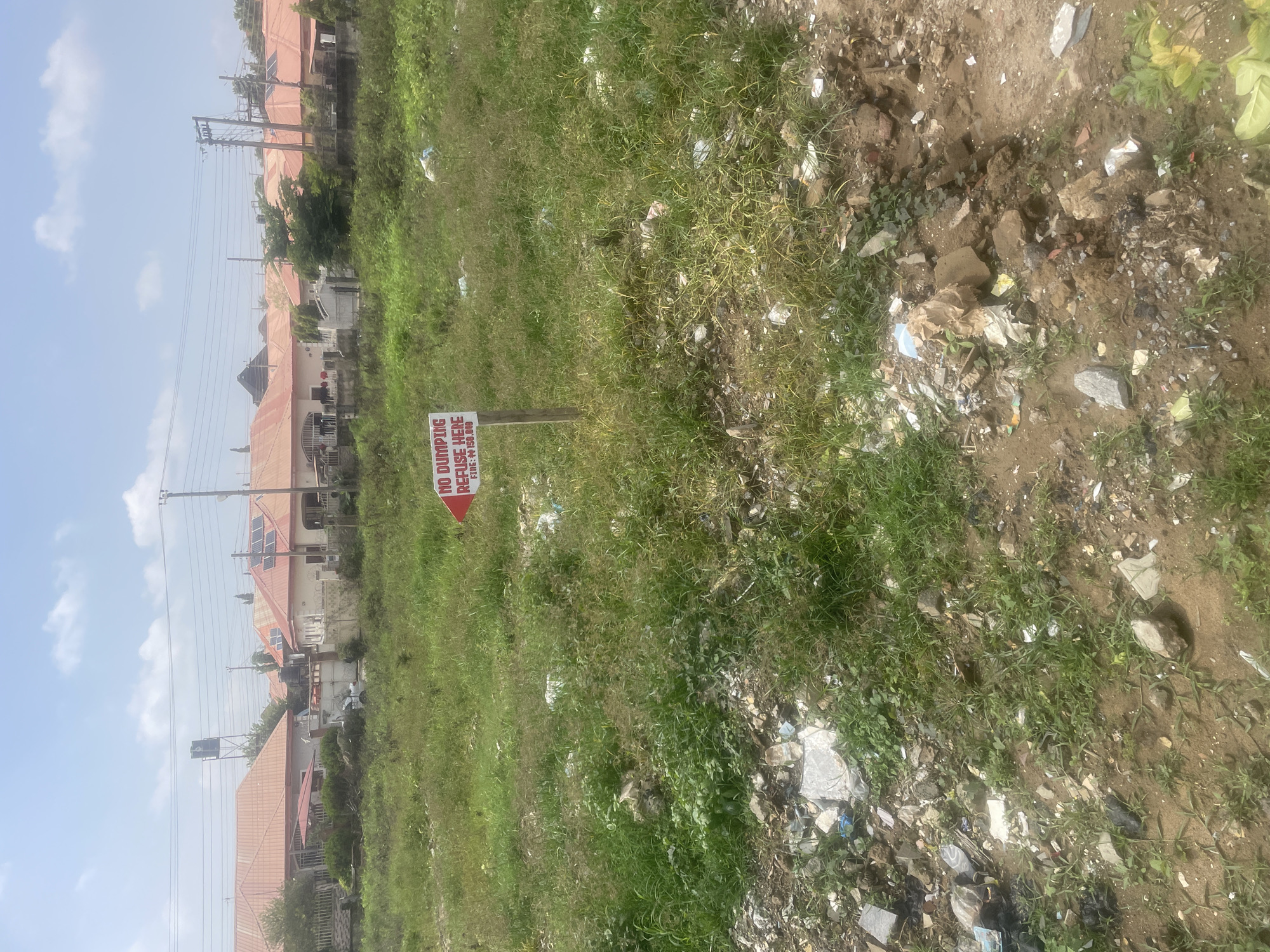 The image captures a partially overgrown, grassy area with scattered debris, likely on the outskirts of a residential neighborhood in Abuja, Nigeria. A red and white sign on a wooden post is prominently placed in the foreground. The sign reads "NO DUMPING REFUSE HERE" in bold black letters, followed by "FINE: N150,000".
Behind the sign, a line of houses with red tiled roofs stretches along a street, suggesting a residential setting. Power lines and poles are visible, indicating the presence of infrastructure. The sky is partly cloudy, and the lighting suggests it is daytime. The overall scene conveys a sense of neglect due to the presence of refuse and overgrown vegetation, contrasting with the nearby modern housing. The sign serves as a clear indication of an environmental concern in the area.