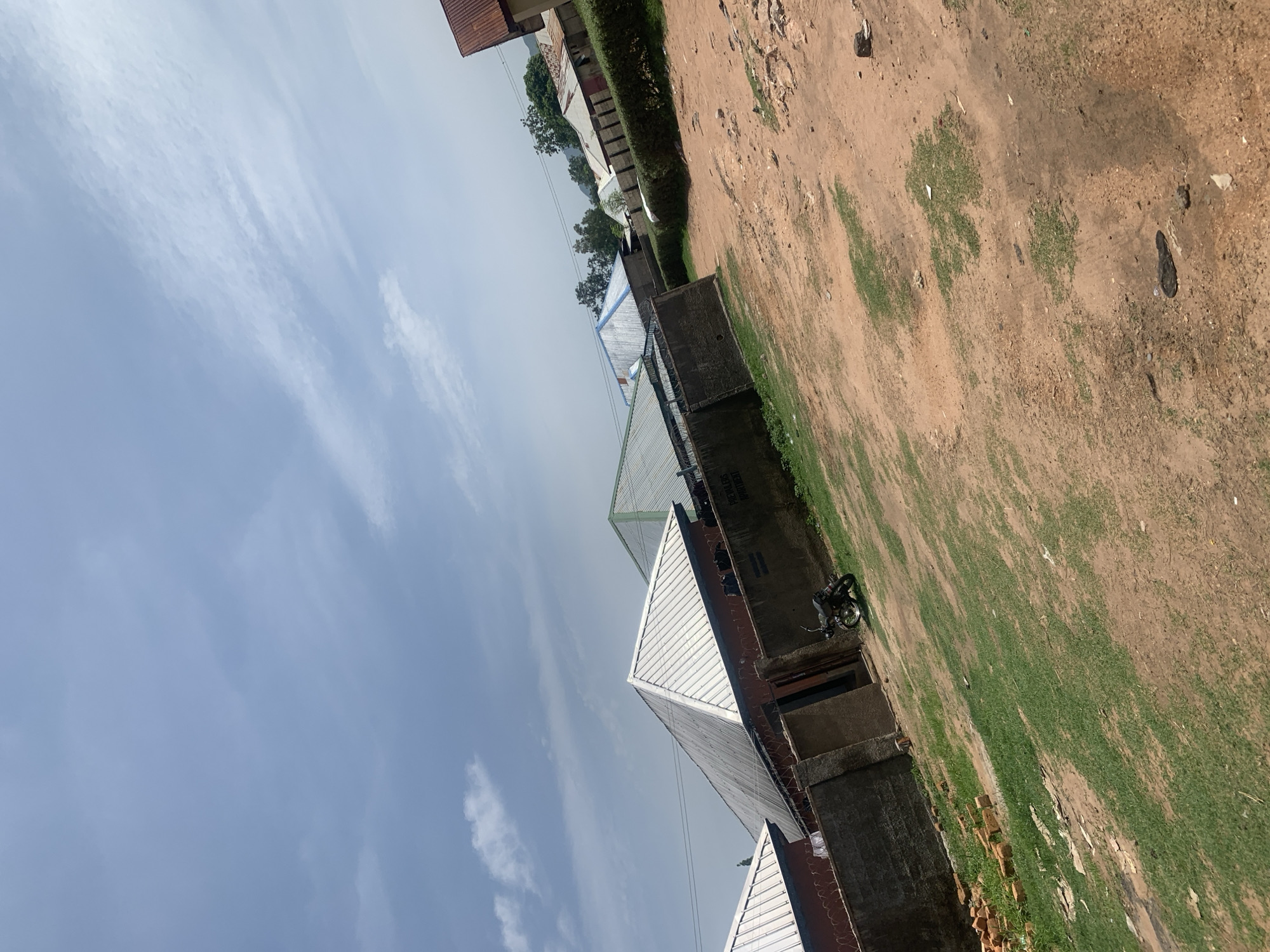 The image captures a view of buildings and a dirt road in Jauro Yinu, Nigeria. The foreground is dominated by a reddish-brown dirt path with patches of green grass and scattered debris. To the left, a pile of bricks sits on the grass. A motorcycle is parked on the dirt path near a concrete wall. Behind the wall, several buildings with metallic roofs are visible, including houses and possibly other structures. The sky above is partly cloudy with a mix of blue sky and white clouds. In the background, more buildings and some trees can be seen, suggesting a residential or semi-rural area within the city. The overall impression is of a somewhat unpaved and sparsely developed urban environment.