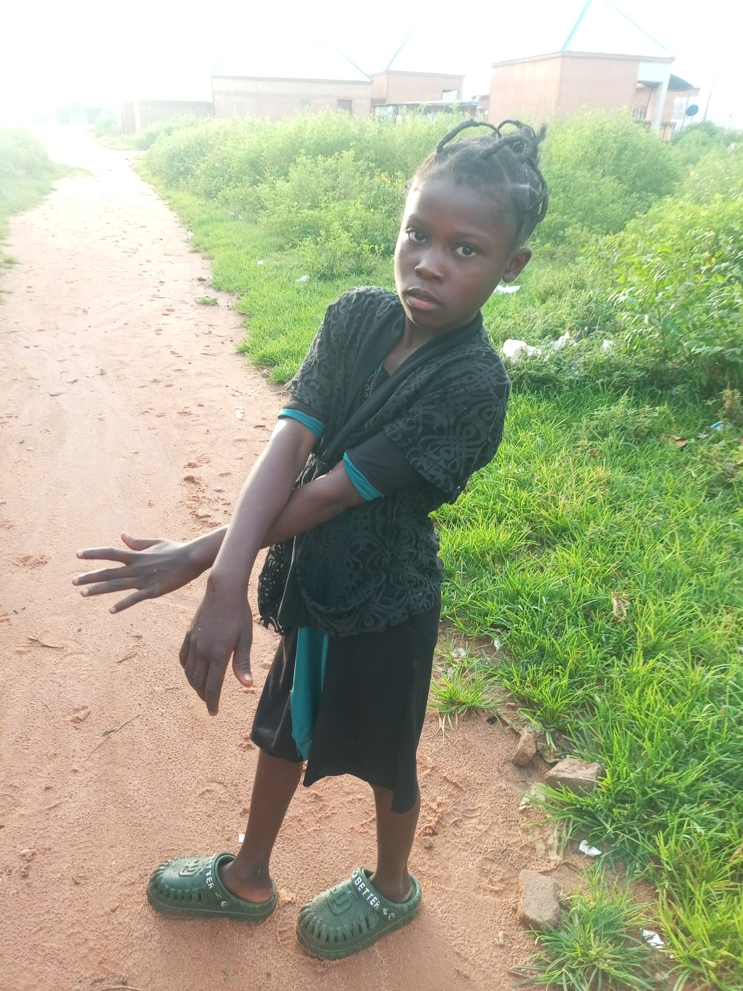 In the city of Tempure, Nigeria, a young child stands on a dirt path beside a grassy verge. The child, with dark skin and braided hair, wears a black short-sleeved top with a patterned overlay and teal trim, and black shorts. Green clogs are on their feet. The child's left arm is bent across their chest, while their right arm is extended outwards with fingers splayed, in a gesture that appears defensive or illustrative. Behind the child, the dirt path continues, flanked by overgrown green grass and scattered debris. In the background, buildings with light-colored walls and peaked roofs are visible, suggesting a residential or developing area. The lighting indicates it is daytime, with a bright, hazy sky.