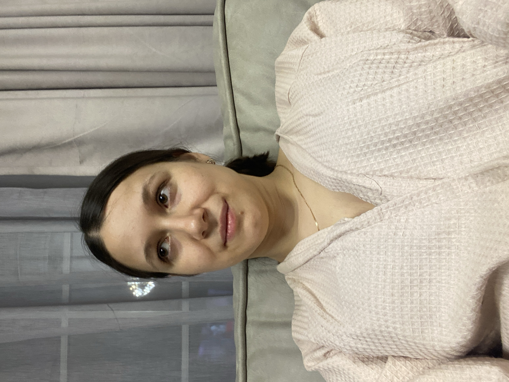 A young woman with dark hair and brown eyes is posing for a selfie. She is wearing a light pink robe and a gold necklace. She is sitting on a gray couch, and behind her is a window with gray curtains. The location in the city of Chaykovsky, Russia is unknown.