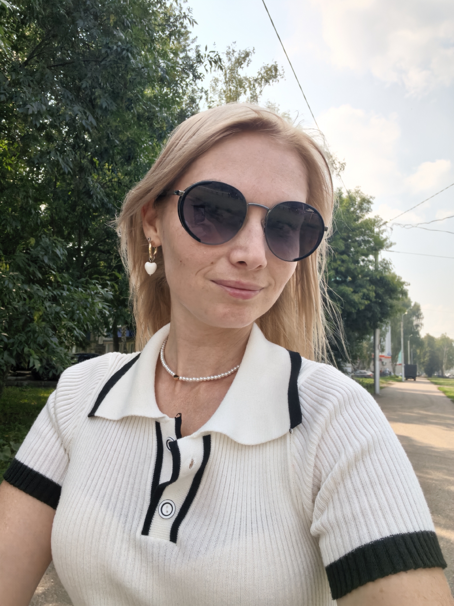 A woman with blonde hair and sunglasses takes a selfie outdoors. She is wearing a white collared shirt with black trim and buttons, a pearl necklace, and heart-shaped earrings. The background shows trees, a sidewalk, and a street with some buildings and vehicles, suggesting an urban or suburban environment. The sky is partly cloudy. The location is the city of Sterlitamak, Russia.