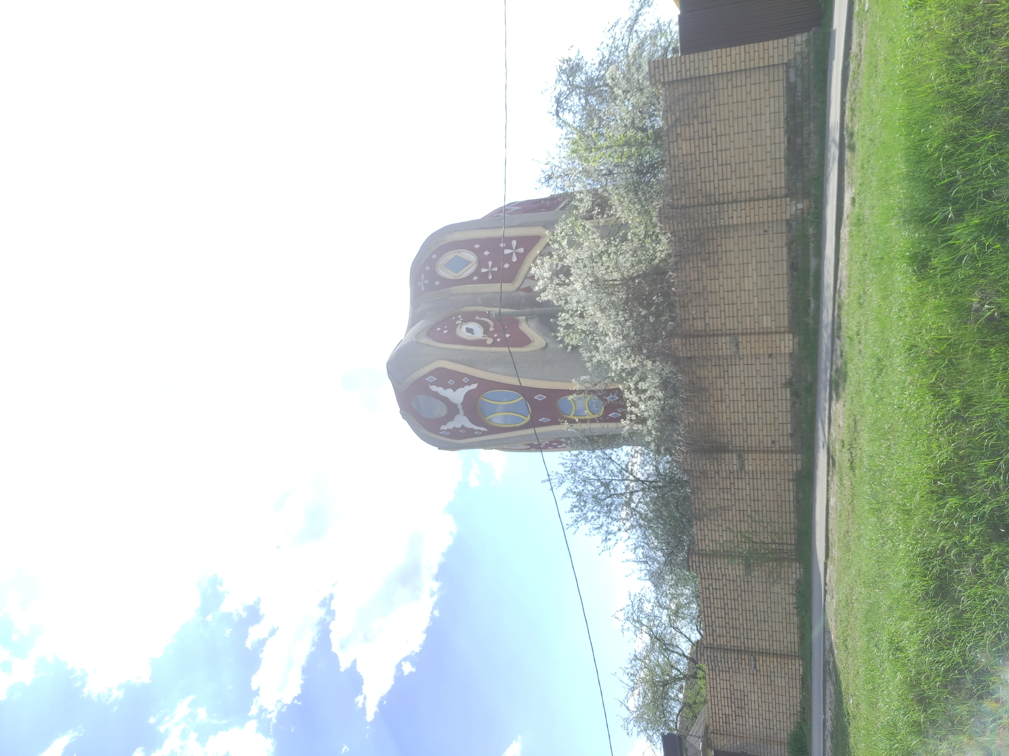 The image shows a decorative monument in Ostrovtsy, Russia, likely a landmark or memorial of local significance. The monument, positioned near a brick fence and grass lawn, features distinct ornamental designs in shades of red, blue, and gold against a light grey background. The sky behind is partially obscured by bright sunlight and scattered clouds. A power line cuts across the frame, running from the monument towards the top right of the image. Trees with white blossoms partially cover the brick fence.