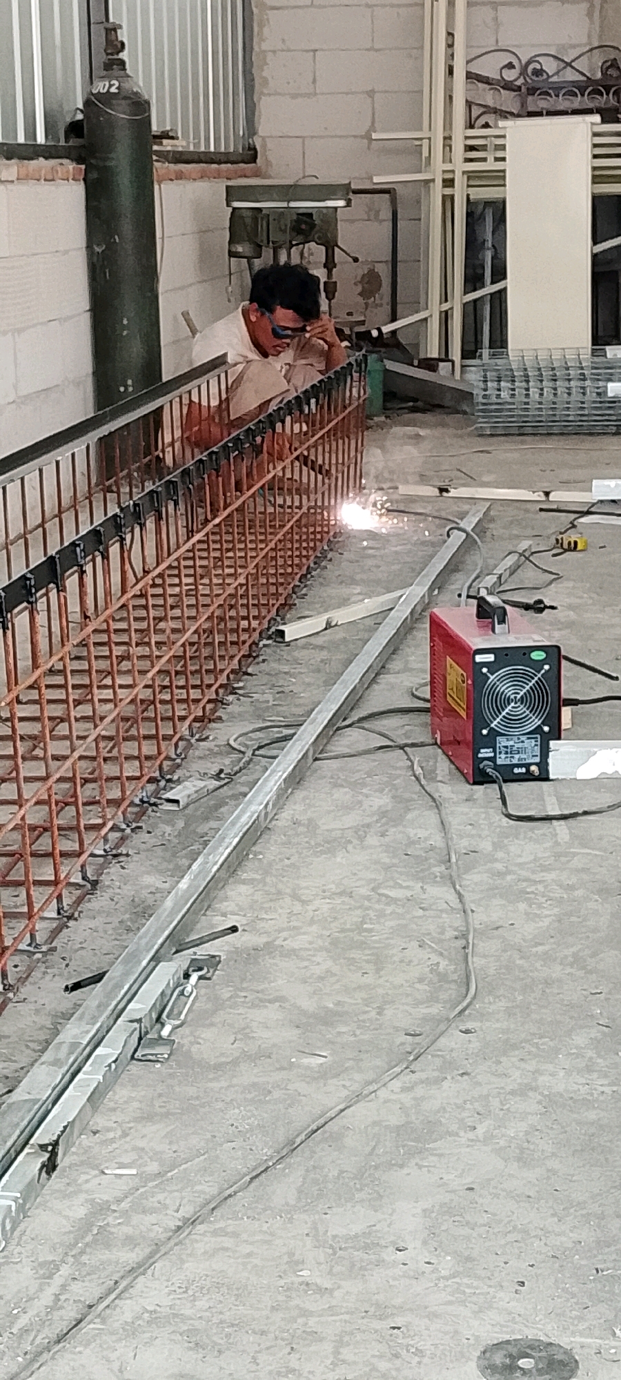 In Sriamur, Indonesia, a welder wearing a light-colored shirt and beige pants is actively engaged in welding a steel reinforcement bar cage. The welder is positioned in the center of the frame, with their head down, focusing intently on the task at hand. Protective eyewear is visible, shielding their eyes from the bright arc. Sparks are visibly flying from the welding point, illuminating the immediate area.
The environment appears to be an industrial or construction site. To the left, a tall, green gas cylinder stands upright, its metallic surface reflecting the ambient light. Behind the welder, a bench drill press is situated against a white brick wall, suggesting a workshop or fabrication area. In the foreground and extending towards the right, a red welding machine with a black fan grille is placed on the concrete floor, connected by cables.
The steel cage being worked on is constructed from numerous thin, reddish-brown bars, arranged in a grid-like pattern. These bars are being joined together by the welder, forming a sturdy structure. Several metal beams, likely for construction purposes, are laid out on the concrete floor around the work area. The ground is made of rough concrete, with some debris and scattered items visible. The overall scene conveys a sense of active construction and manufacturing.