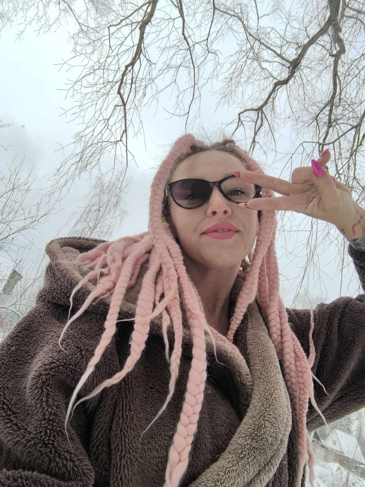 The image depicts a person in what appears to be a snowy outdoor environment. The individual is wearing sunglasses, pink lipstick, and pink braids. They are also wearing a brown, furry hooded jacket. The person is holding up a hand in a "peace" or "victory" sign. Bare tree branches are visible in the background. Based on the context provided, the location is Noviy, Russia. It is a low-angle shot, with the camera pointed upwards.
