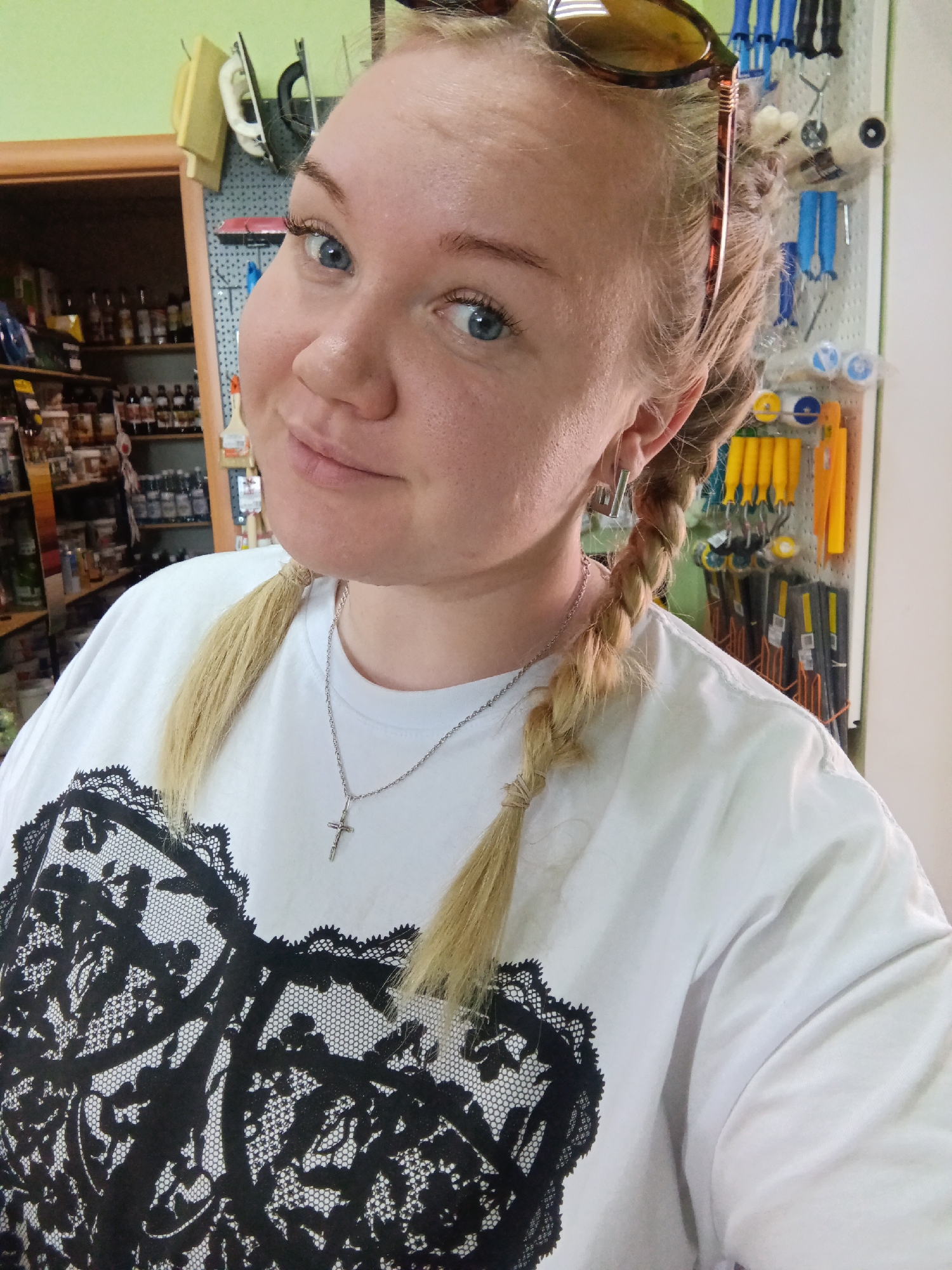 A blonde-haired woman with blue eyes is posing for a selfie. She is wearing her hair in two braids and has sunglasses perched on top of her head. She is wearing a white t-shirt with a black lace design on the front and a silver necklace with a cross pendant. The background appears to be a hardware store or a workshop. Shelves stocked with various items and tools are visible. The city of Фоки, Russia is not recognizable in this image.