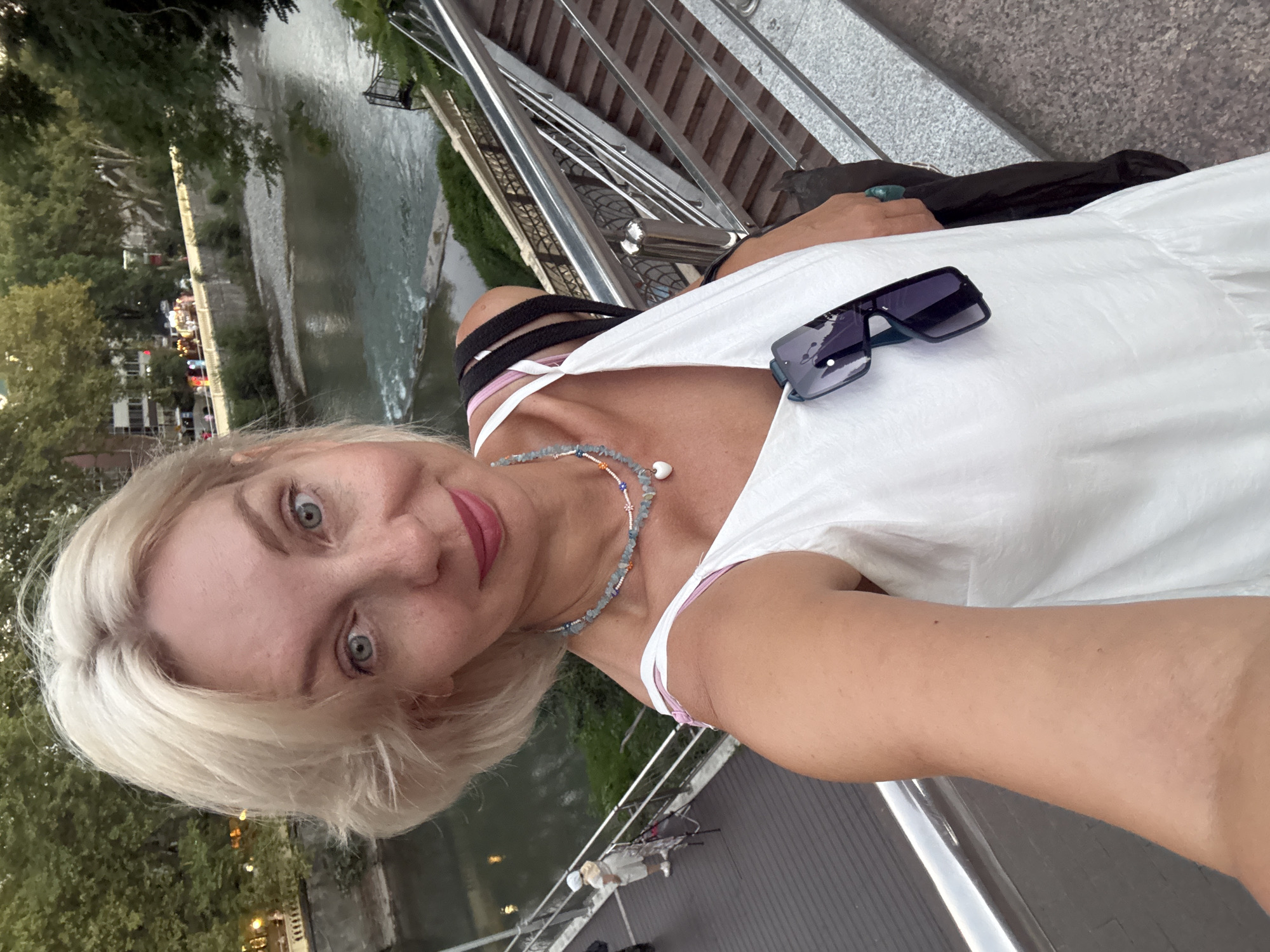 An outdoor, close-up, selfie-style photograph shows a woman with blonde hair and blue eyes, wearing a white sleeveless dress and a beaded necklace with a heart-shaped pendant. Sunglasses are tucked into the neckline of her dress. She is standing on a walkway with a metal railing, adjacent to a set of stairs, with a river and trees in the background. The scene appears to be along the Sochi River embankment in Sochi, Russia. The lighting suggests it is late afternoon or early evening.