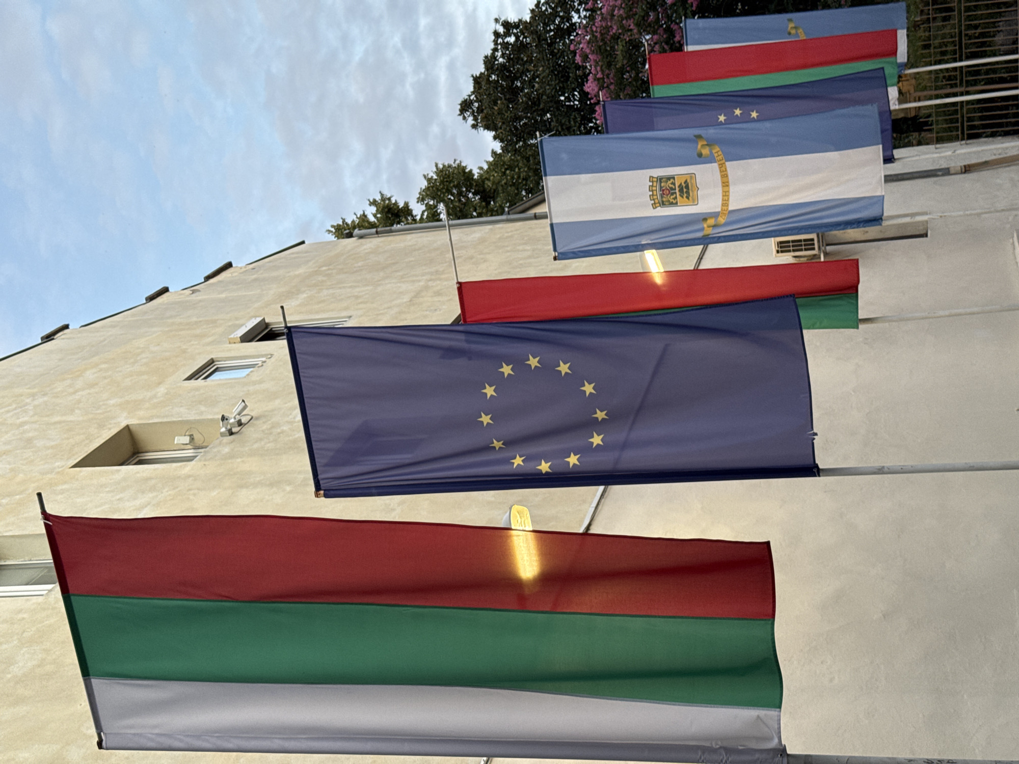 The image shows several flags displayed vertically along the exterior wall of a building. The building has a beige facade with a few windows and an air conditioning unit visible. The sky is cloudy and overcast.
The flags from left to right are:
1. A tricolor flag with vertical stripes of white, green, and red, which is the flag of Bulgaria.
2. A dark blue flag with a circle of yellow stars, which is the flag of the European Union.
3. A flag with a coat of arms and text, featuring vertical stripes of light blue and white. The text appears to be in Cyrillic. This is likely the flag of a municipality within Bulgaria.
4. A dark blue flag with yellow stars.
5. A tricolor flag with vertical stripes of green, red, and white, which is the flag of Bulgaria.
The arrangement of these flags suggests a display of national and possibly regional or institutional symbols. Based on the presence of the Bulgarian flag and the specific municipal flag, the location is likely within Bulgaria. Without further information about the coat of arms on the municipal flag, it's difficult to pinpoint the exact location. However, the style of the building and the overall scene are consistent with an urban environment in Bulgaria.