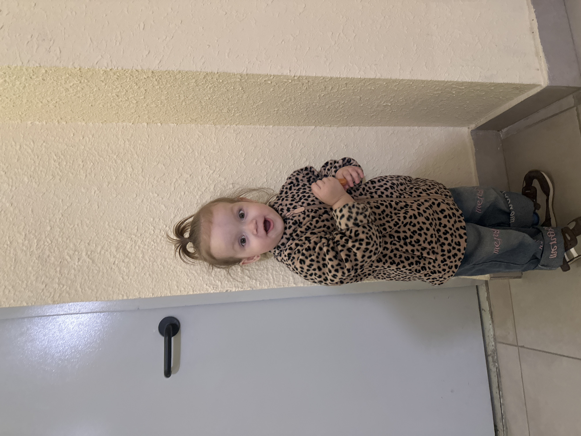 An indoor, eye-level full-length shot depicts a young child, approximately two to three years old, standing in what appears to be an interior hallway in Yekaterinburg, Russia. The child, with light brown hair pulled into a high bun, faces forward with wide eyes and an open mouth, exhibiting an animated expression. They are dressed in a long-sleeved fleece top featuring a black and tan leopard print, blue denim jeans with pink embroidered text (partially visible as "mery" and "wow") on the lower legs, and brown and white shoes. The child holds a small, orange, cylindrical object in their clasped hands. To the left of the child is a light grey door equipped with a horizontal black door handle. To the child's right and extending behind them is a light beige-yellow wall with a heavily textured finish, featuring a distinct vertical band of slightly varied texture or color. A grey baseboard runs along the bottom of both the wall and the door frame. The floor is composed of light-colored square tiles with visible grout lines.