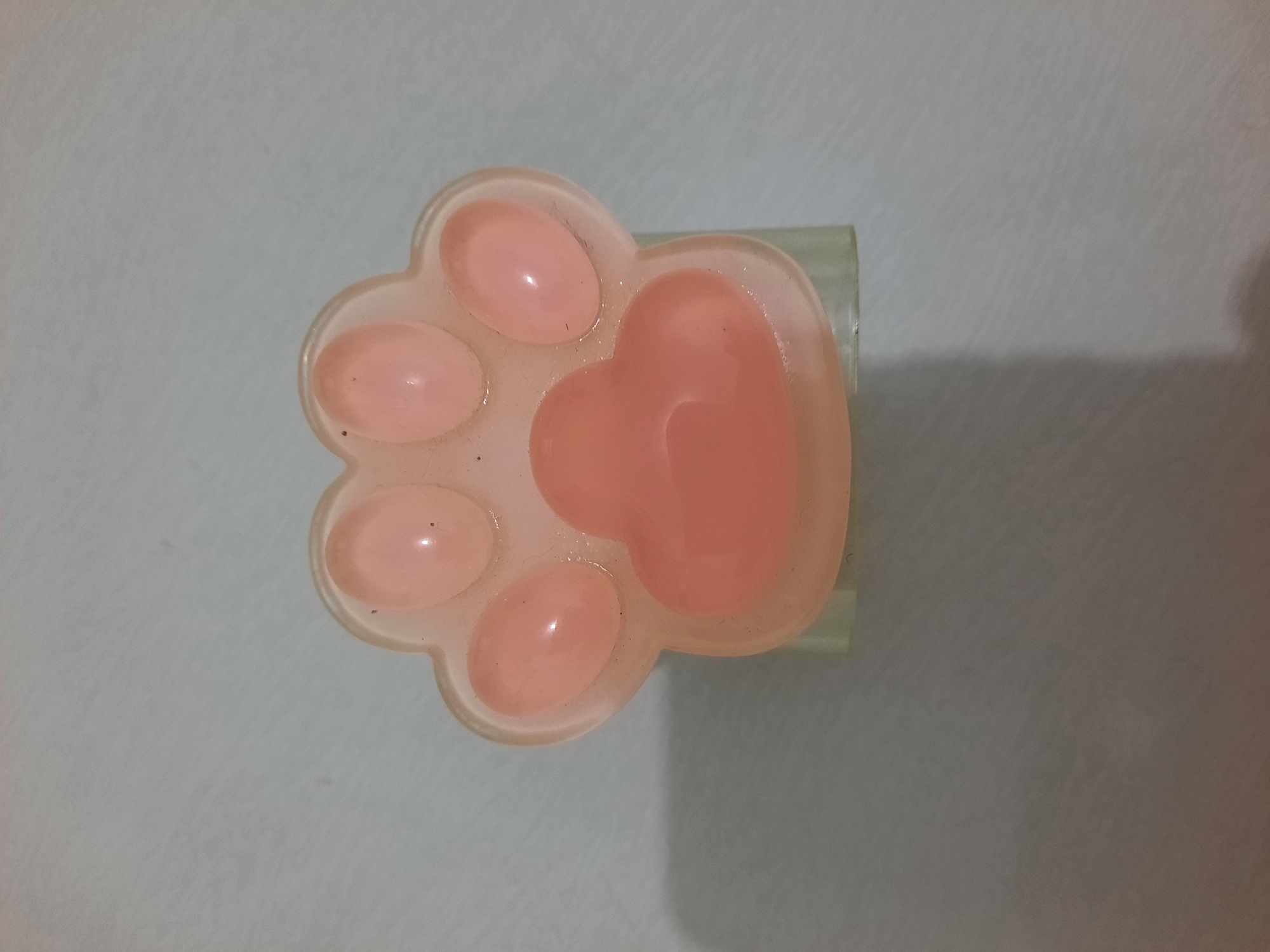 A light pink paw-shaped object, possibly a stress toy or bath bomb, rests on a pale blue textured surface. The object has four circular indentations at the top, representing toes, and a larger heart-shaped indentation at the bottom, representing the palm pad. The surface appears to be a tabletop or counter. The lighting creates a soft shadow to the left of the object. There are no people or other objects present. The location is not specified within the image.