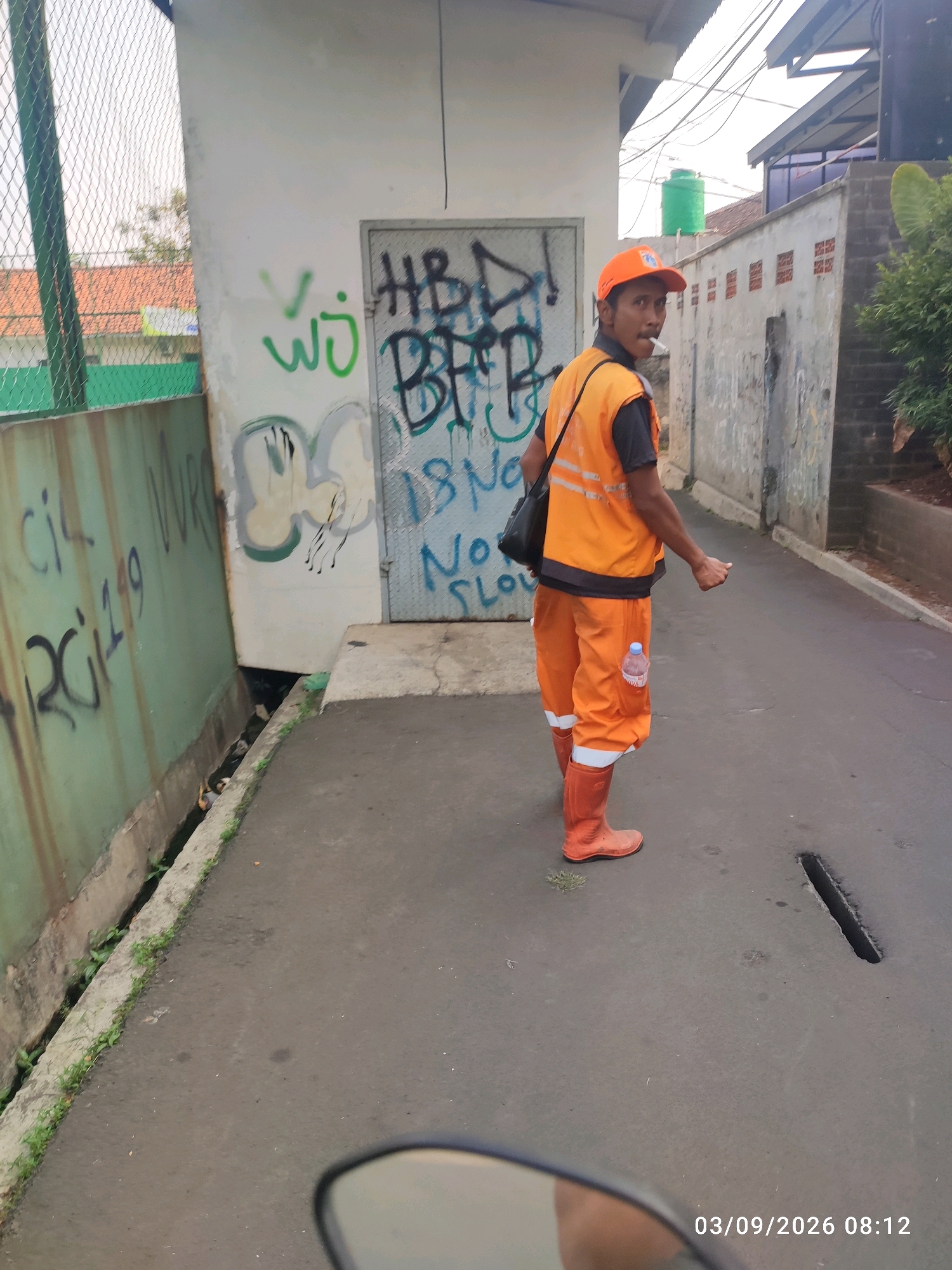 The image depicts a street scene in East Jakarta, Indonesia. In the foreground, a person wearing an orange high-visibility vest, orange pants, and orange boots is visible. The person is holding a cigarette in their mouth and looking towards the camera. A black bag is slung over their shoulder. The background shows a building with graffiti on its walls. There is a fence to the left of the building, and some greenery is visible. The ground is paved with asphalt. The date and time stamp "03/09/2026 08:12" is present in the bottom right corner of the image.