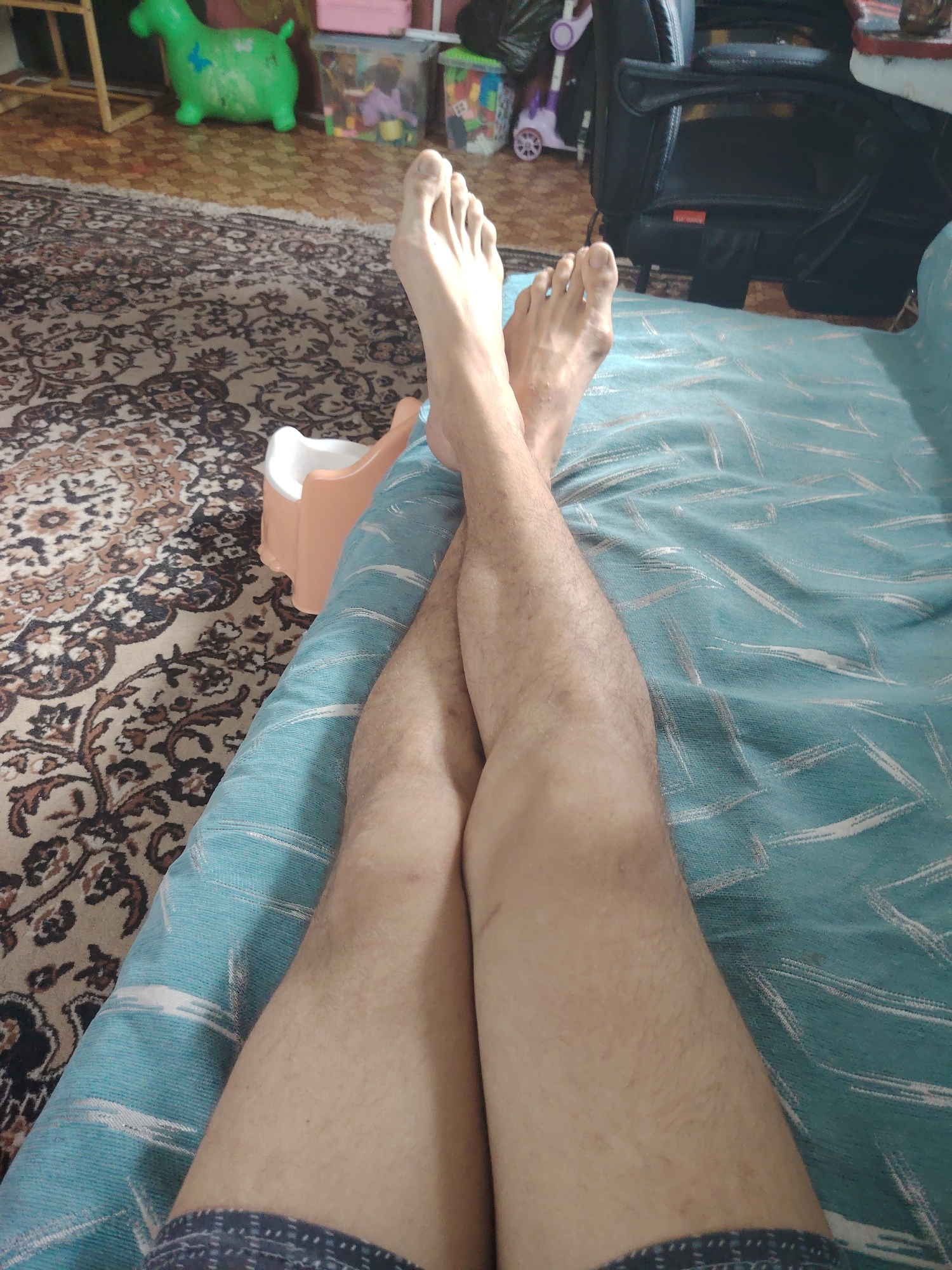 The image depicts a person's legs crossed, resting on a turquoise-colored blanket with a white abstract pattern. The legs appear to be bare from the knees down. The left leg is closer to the viewer and has a bandage or wrap around the lower calf and ankle. The right leg is positioned on top of the left. In the background, a patterned rug covers the floor. Several objects are visible on the rug, including a small, light-colored potty chair, children's toys, and a portion of a black office chair. The lighting suggests an indoor setting. The geographical location of Tokmok City, Kyrgyzstan, is not discernible from the visual information present in the image.