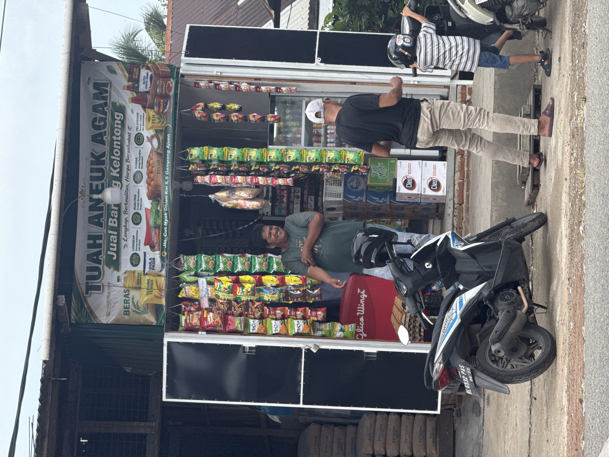 An exterior street-level, eye-level shot depicts a small, open-air convenience store, likely a "warung," in the city of Ajuen, Indonesia. A large, horizontal banner above the store entrance reads "TUAH ANEUK AGAM Jual B..." and "g Kelontong," indicating it sells various goods. The store itself is filled with shelves displaying numerous bagged snacks, beverages, and packaged food items.
In the foreground, a black and white scooter is parked. A man with dark hair and wearing a green t-shirt stands to the right of the scooter, facing the store and smiling. To the right of him, another man wearing a white baseball cap and a black t-shirt is facing away from the camera, seemingly interacting with the store's contents. A young boy in a striped shirt and shorts stands to the far right, holding onto the handlebars of a motorcycle. The ground surface is a mix of concrete and dirt. Stacked sacks of what appear to be cement or building materials are visible to the left of the store. The environment suggests a moderately busy, possibly humid, day with the store operating on a street.