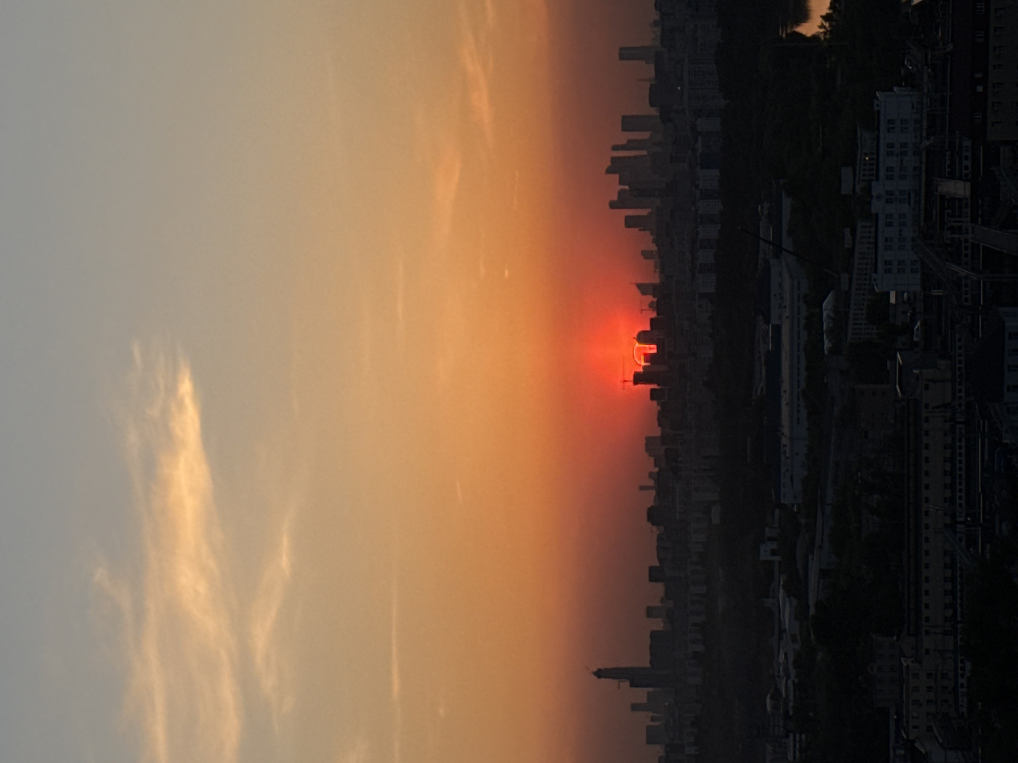 The image depicts a sunset over Moscow, Russia. The sky transitions from a hazy gray-blue on the left to a vibrant orange and red where the sun is setting, partially obscured by the city skyline. The skyline is silhouetted against the sunset, showing a mix of high-rise buildings and lower structures. Details within the cityscape are hard to discern due to the darkness and distance, but individual buildings can be made out. The horizon line, where the sky meets the city, is not perfectly straight. There are some trees visible amid the buildings. The overall impression is one of an urban sunset, with the natural beauty of the sky contrasted against the built environment of Moscow.