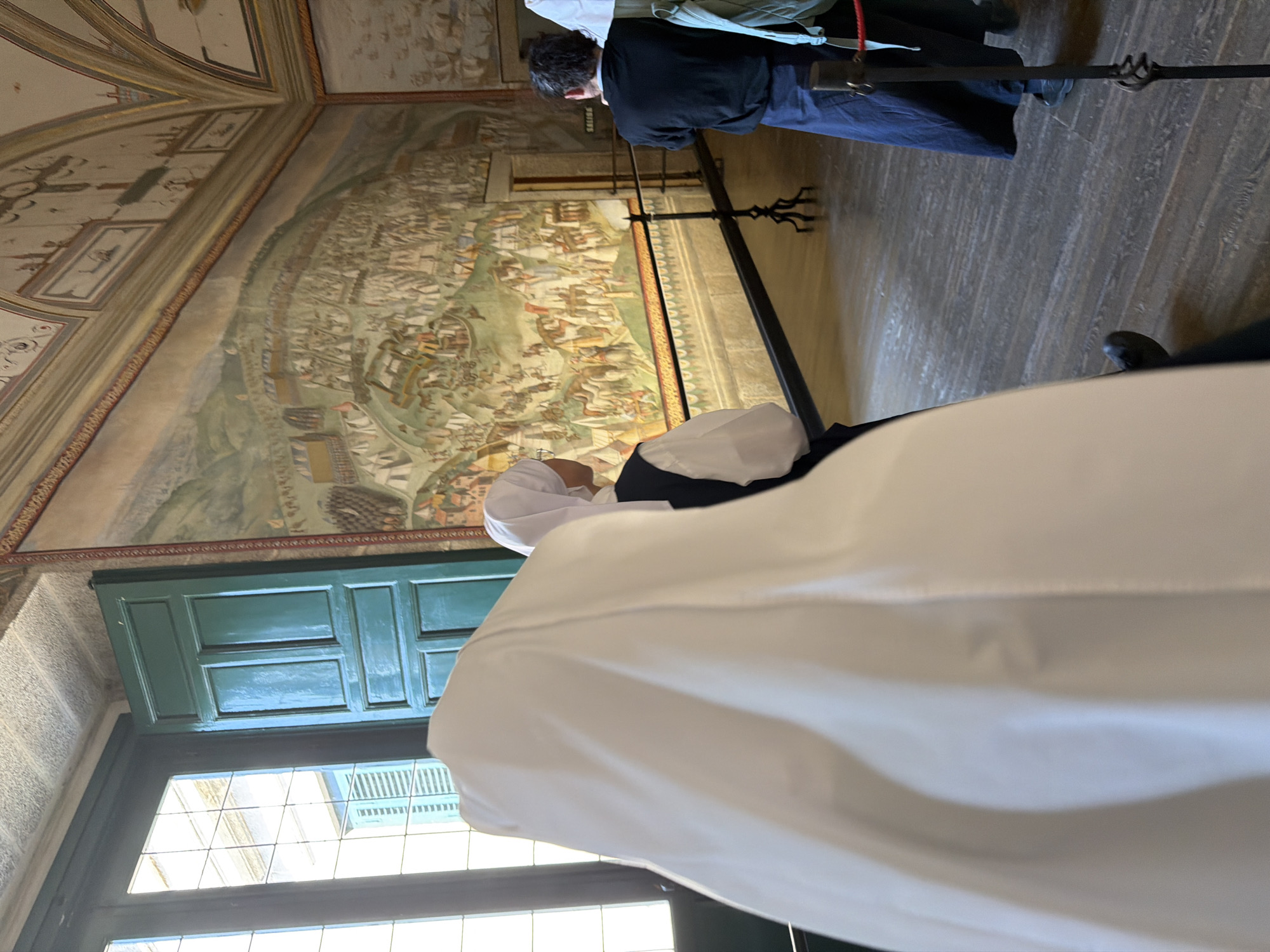 In the Palace of the Escorial, San Lorenzo De El Escorial, Spain, two people dressed as nuns stand in a room with a large mural on the wall. The first nun, in the foreground, is mostly obscured by a large white habit, with only a white wimple visible. The second nun, in the middle ground, wears a white wimple and a dark vest over a white shirt, with her back to the camera. A third person, a man in a black shirt and dark pants, stands further back in the room. The room has a wooden floor and a large mural depicting a historical scene covers the wall. A velvet rope barrier separates the visitors from the mural. A window with green shutters is visible on the left side of the image.
