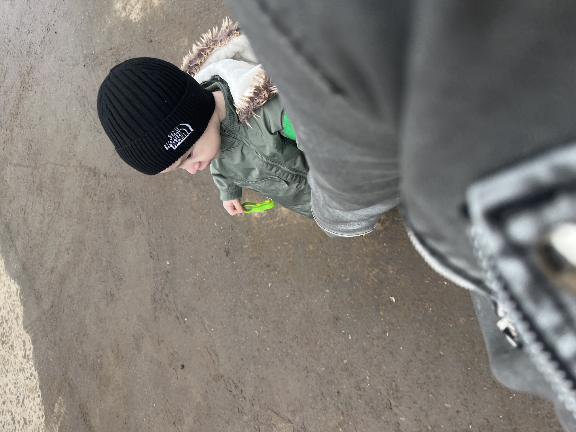 This high-angle, top-down shot captures a young child walking outdoors, likely alongside an adult whose leg and foot are partially visible on the right side of the frame. The scene is set on a damp, earthy path in Chaikovsky, Russia, suggesting cool or possibly wet weather conditions.
The child, appearing to be a toddler or preschooler, is dressed warmly in a black ribbed beanie featuring the distinctive white "THE NORTH FACE" logo. They wear a green hooded jacket with a visible white faux fur lining, indicating appropriate attire for cooler temperatures. In their right hand, the child holds a bright green plastic object, possibly a toy or a small tool. Their head is tilted downwards, and their gaze seems to be fixed on the ground or the object they are holding, suggesting a moment of focus or exploration during their walk.
The adult, from whose perspective the photo is taken, is wearing grey pants or sweatpants and a dark shoe or boot, partially seen in the foreground on the right. The overall impression is a casual outdoor activity, such as a stroll or playtime, on an overcast or damp day.